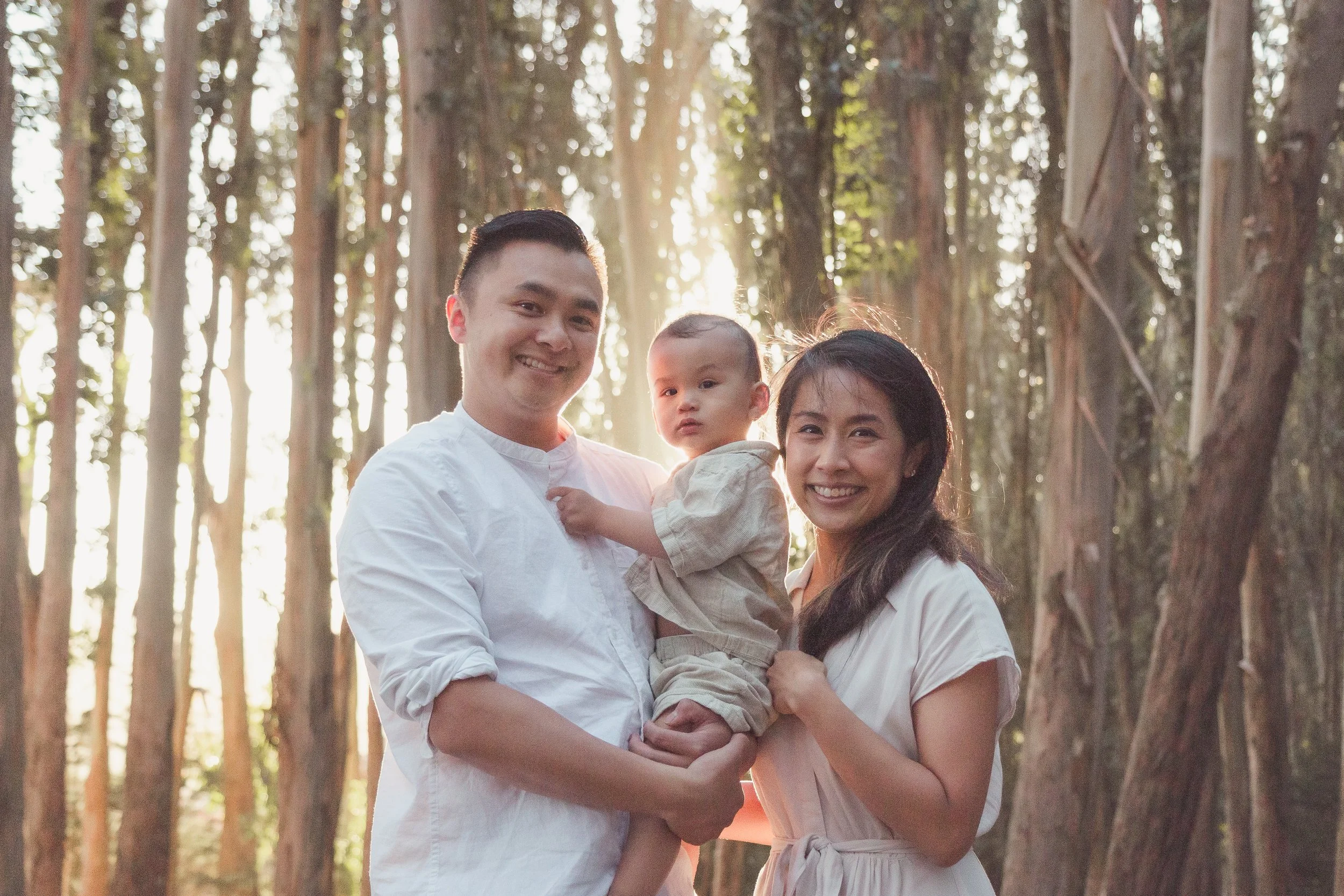 A smiling family of three, a man, a woman, and a young child, standing in a forest with tall trees and sunlight filtering through.