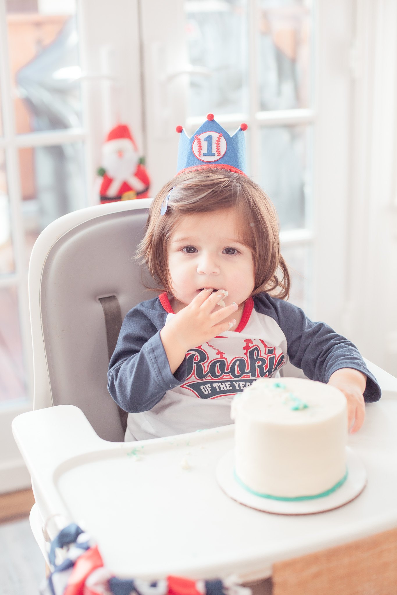 Young girl with brown hair wearing a blue birthday hat marked with a baseball and the number one, celebrating her first birthday at a table with a white cake. She is wearing a baseball-themed shirt with the words "Rookie of the Year" and eating cake