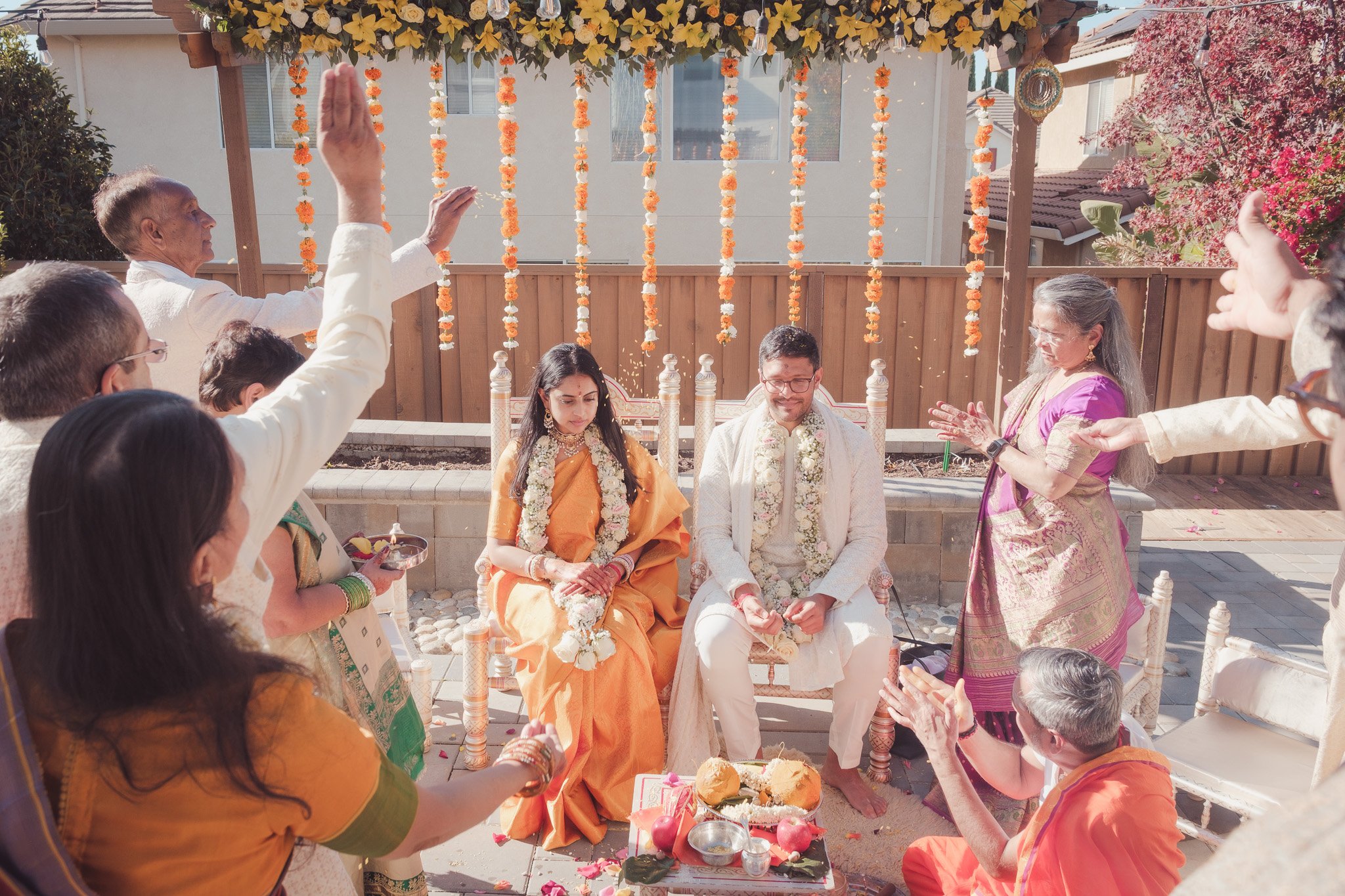 Indian wedding ceremony outdoors with bride and groom seated on a decorated platform, surrounded by family and friends performing prayer rituals.