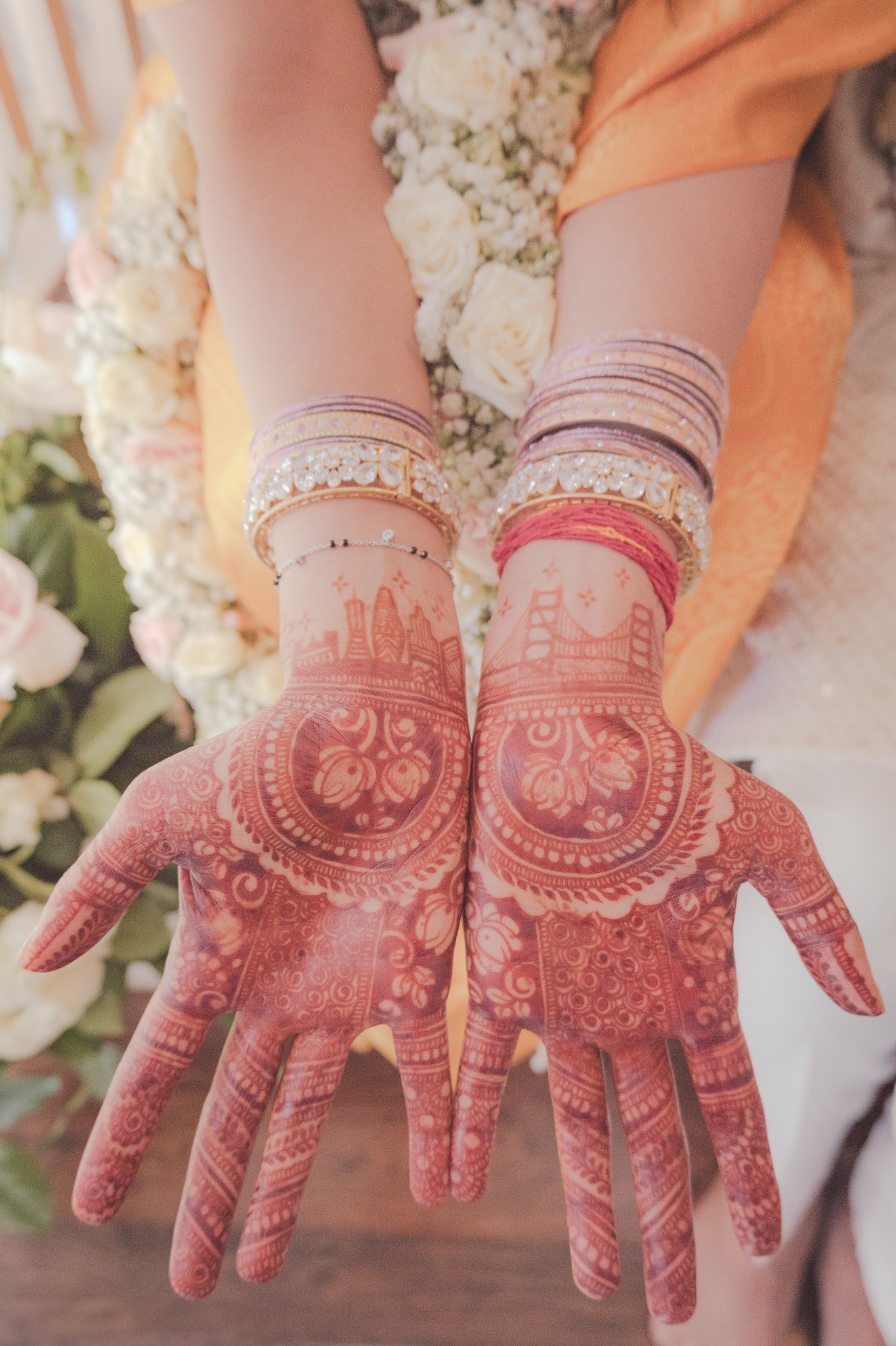 Close-up of hands with intricate henna designs, jewelry, and floral garlands, surrounded by white and pink flowers.