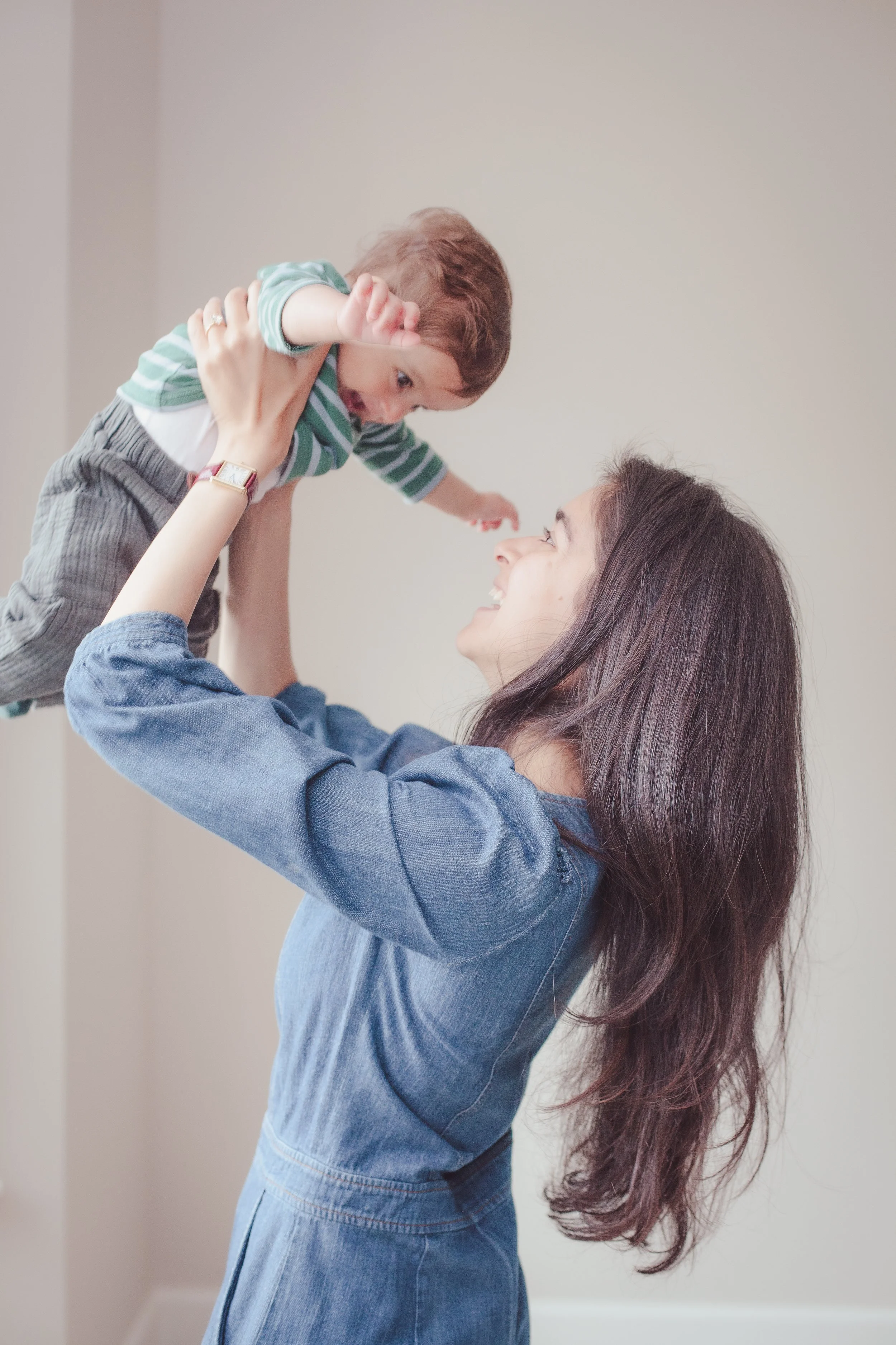 A woman with long dark hair lifts a young boy in the air. The boy is smiling and reaching out with his arms. The woman is smiling and looking up at the boy, both seem happy and playful.