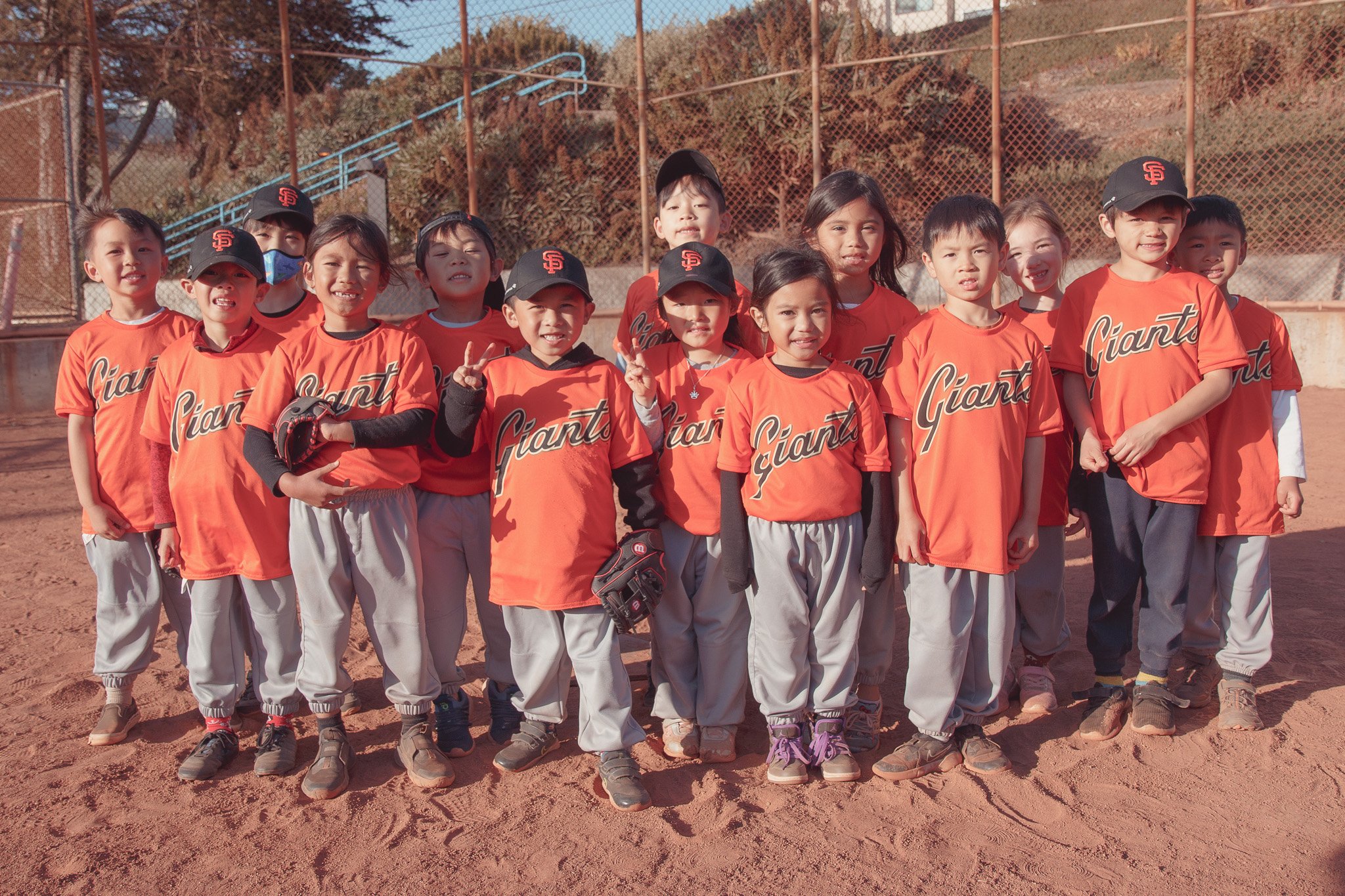 Youth baseball team wearing orange jerseys and gray pants posing on a dirt field, some holding baseball gloves, with a chain-link fence and trees in the background.