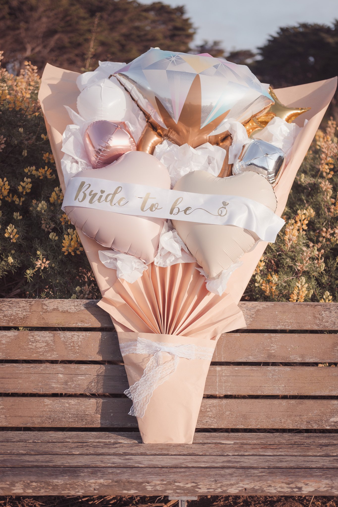 Flower bouquet made of heart-shaped and star-shaped balloons in pastel pink, white, bronze, silver, and gold with a ribbon that reads 'Bride to be' in gold lettering.