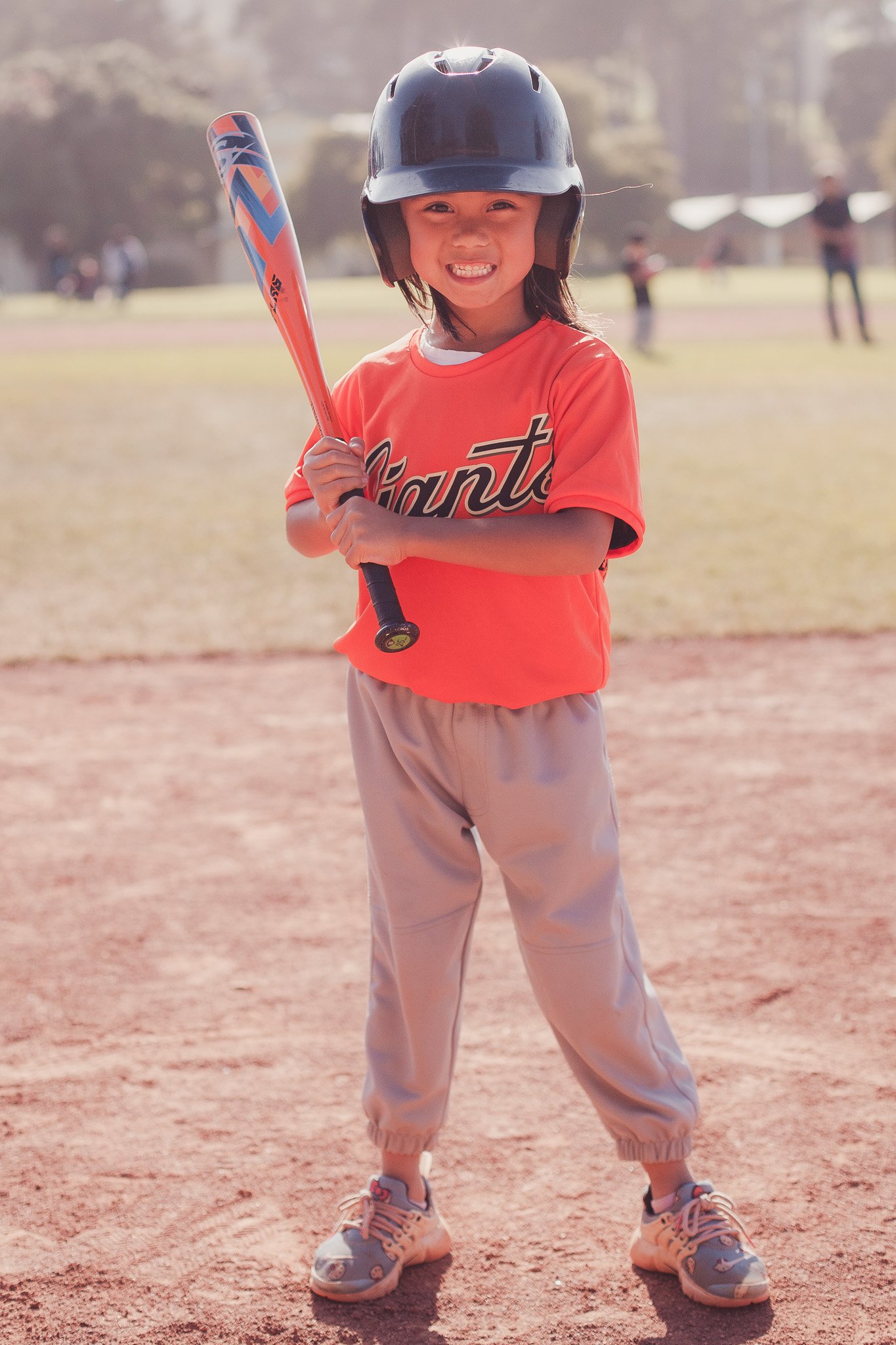 A young girl wearing a black helmet, orange sports jersey with the word 'Santa' on it, beige athletic pants, and pink sneakers, standing on a baseball field with a bat in her hands, smiling at the camera.