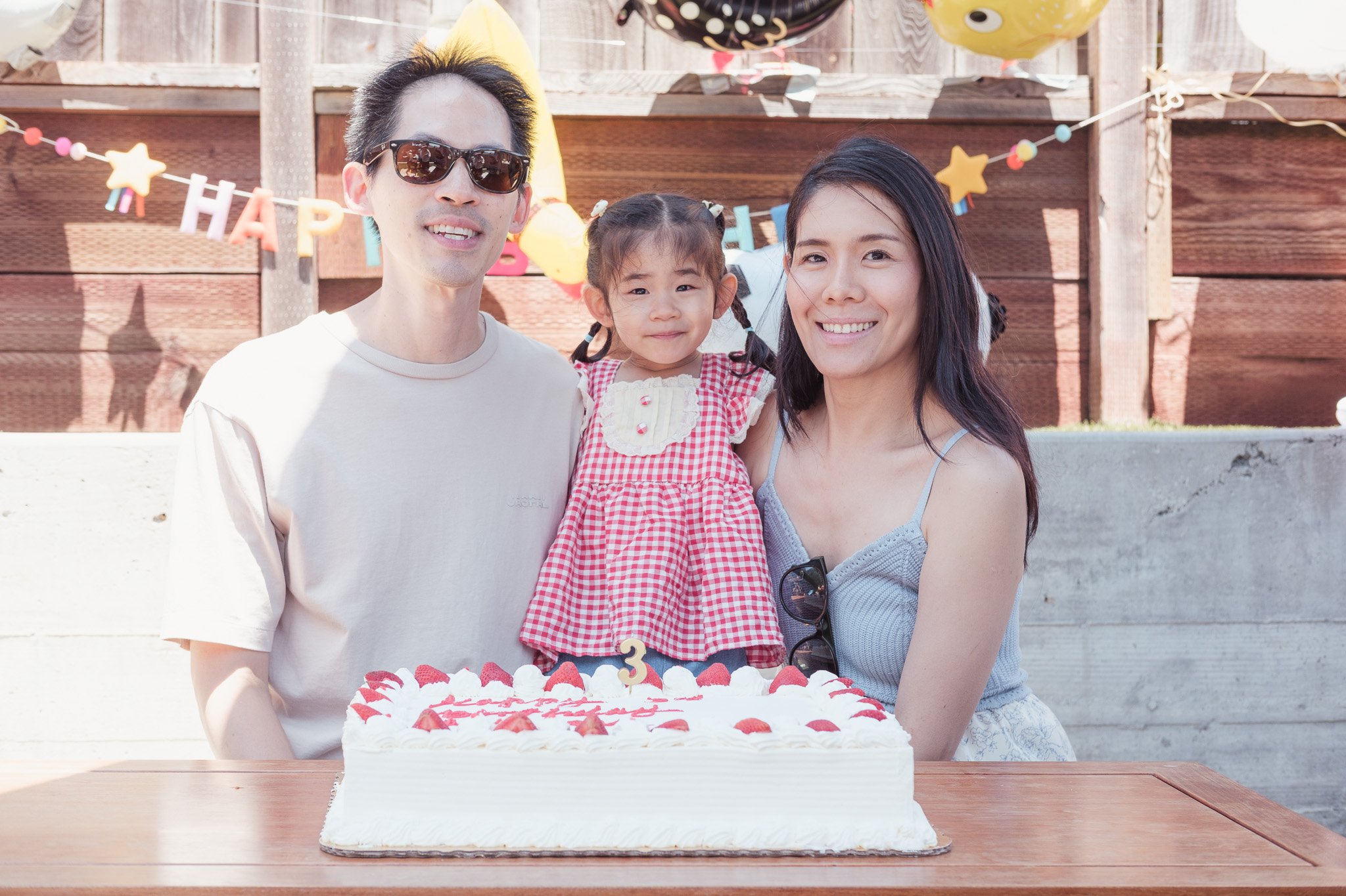 A family celebrating a third birthday, with a young girl in a red and white checkered dress, a cake with strawberries and a number 3 candle, outside in a backyard decorated with balloons and a banner that says 'HAPPY'.