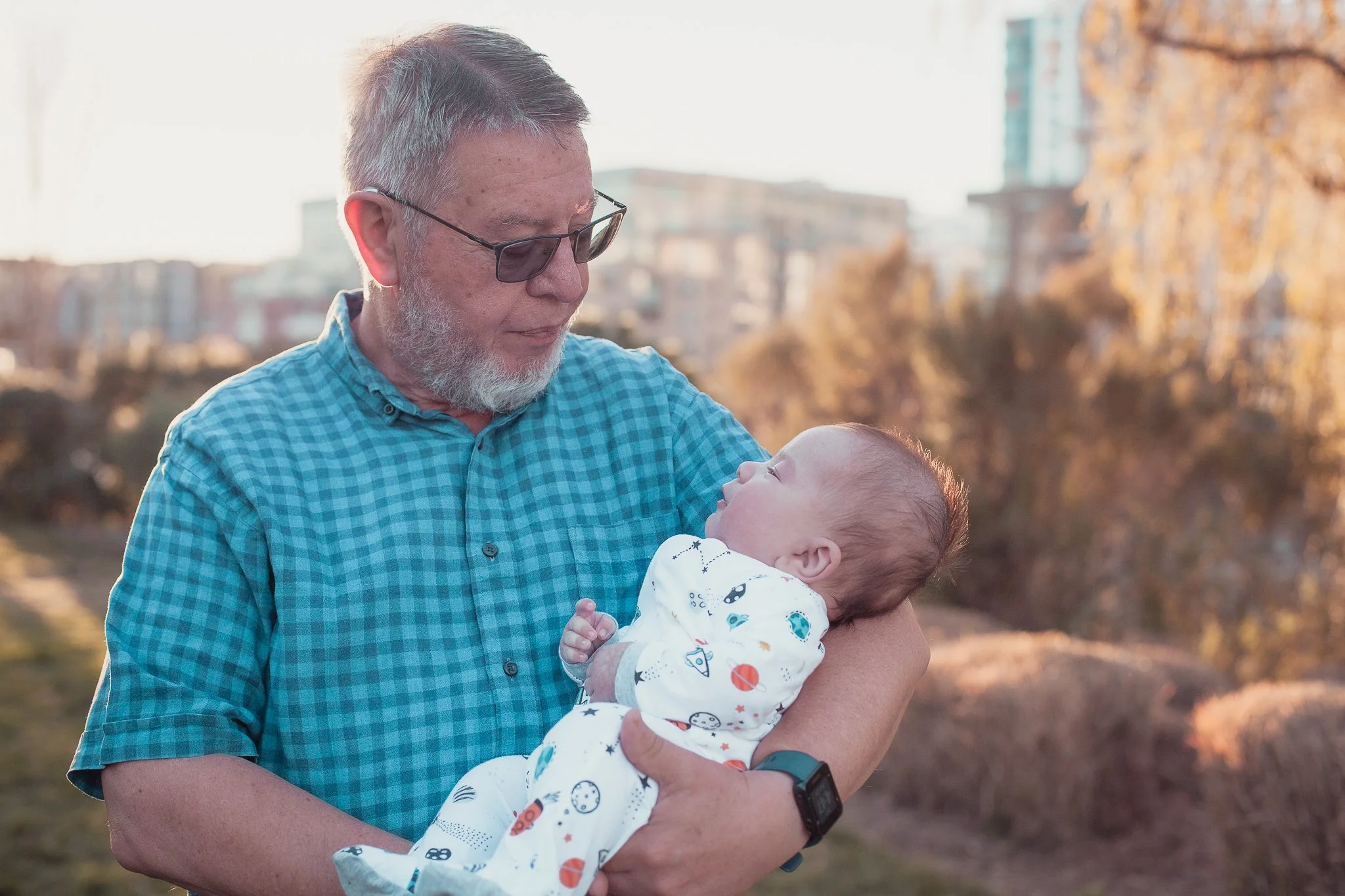 An elderly man with glasses and a gray beard holds a baby in his arms outdoors during sunset, looking at the baby with affection. The baby is dressed in a white outfit with space-themed illustrations and looks up at the man. The background shows trees with autumn leaves and buildings.