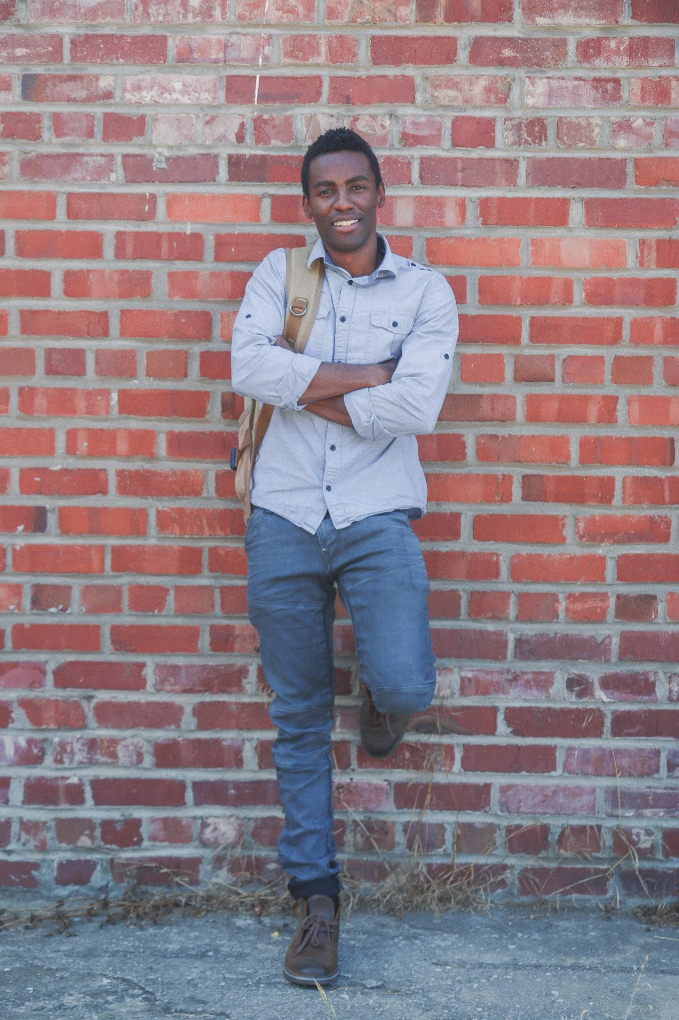 A young man with dark hair, wearing a light gray button-up shirt with rolled-up sleeves, dark jeans, and brown shoes, stands with arms crossed and rests one foot on the wall behind him, leaning against a brick wall with some dried grass at his feet.