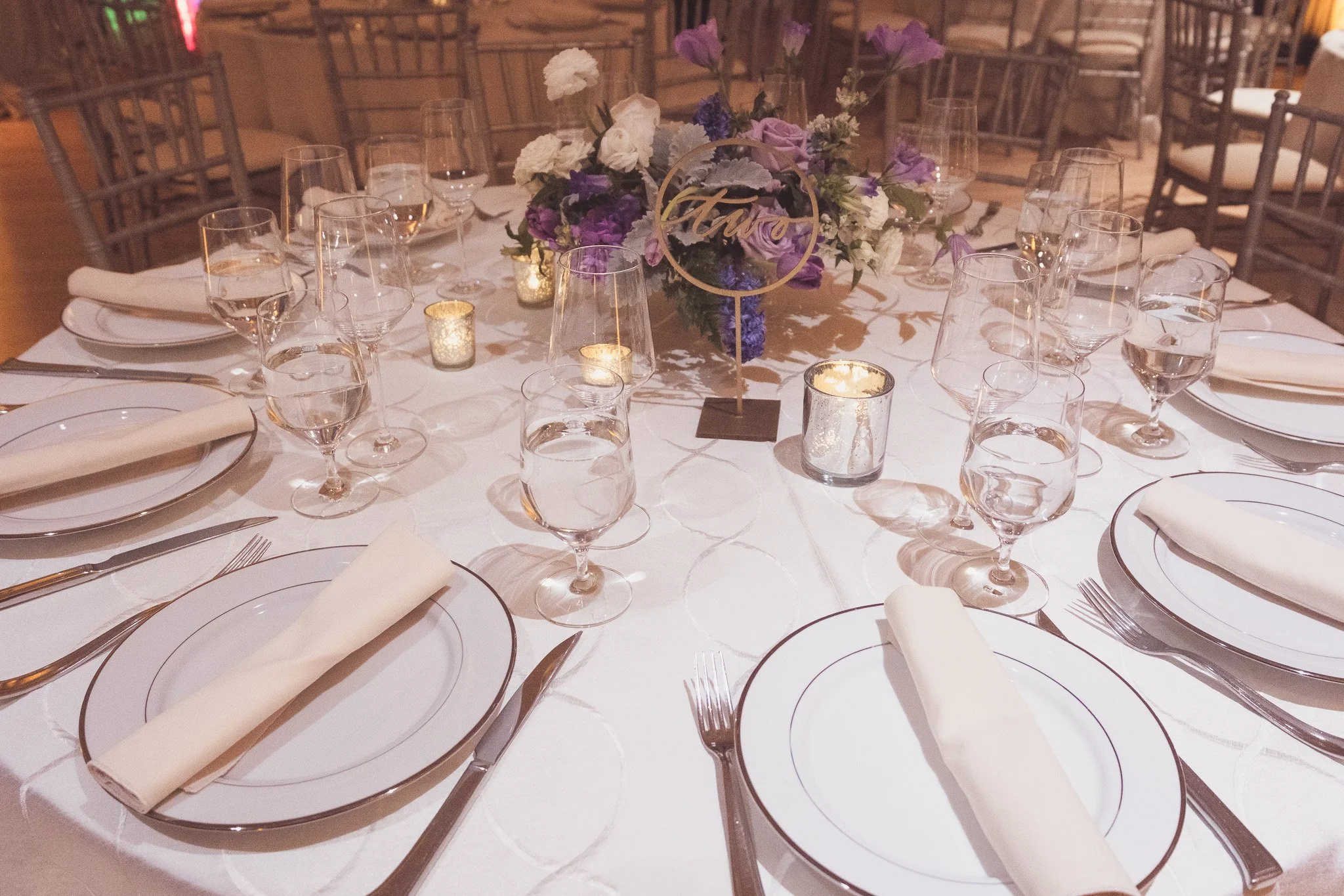 Elegant round wedding or formal dinner table set with white plates, silverware, white napkins, multiple wine glasses, small candles, and a floral centerpiece with white and purple flowers. A decorative sign reading 'Love' is in the center.