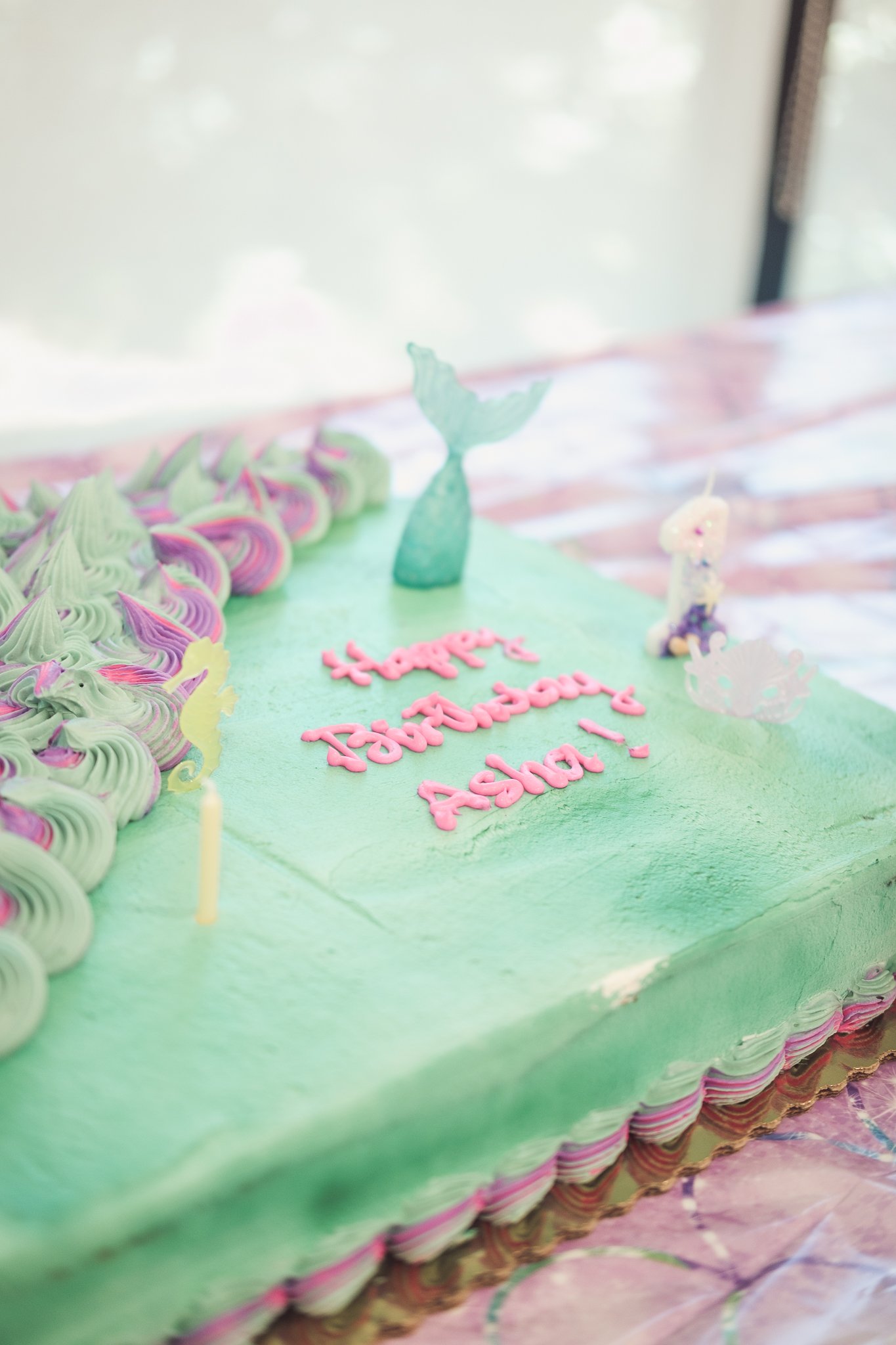 A rectangular ice cream cake with green frosting border and pink and green swirled frosting on top. The cake has pink icing message saying "Happy Tourette's Awareness!" and has decorative sea-themed toppers, including a whale tail and fairy figurines