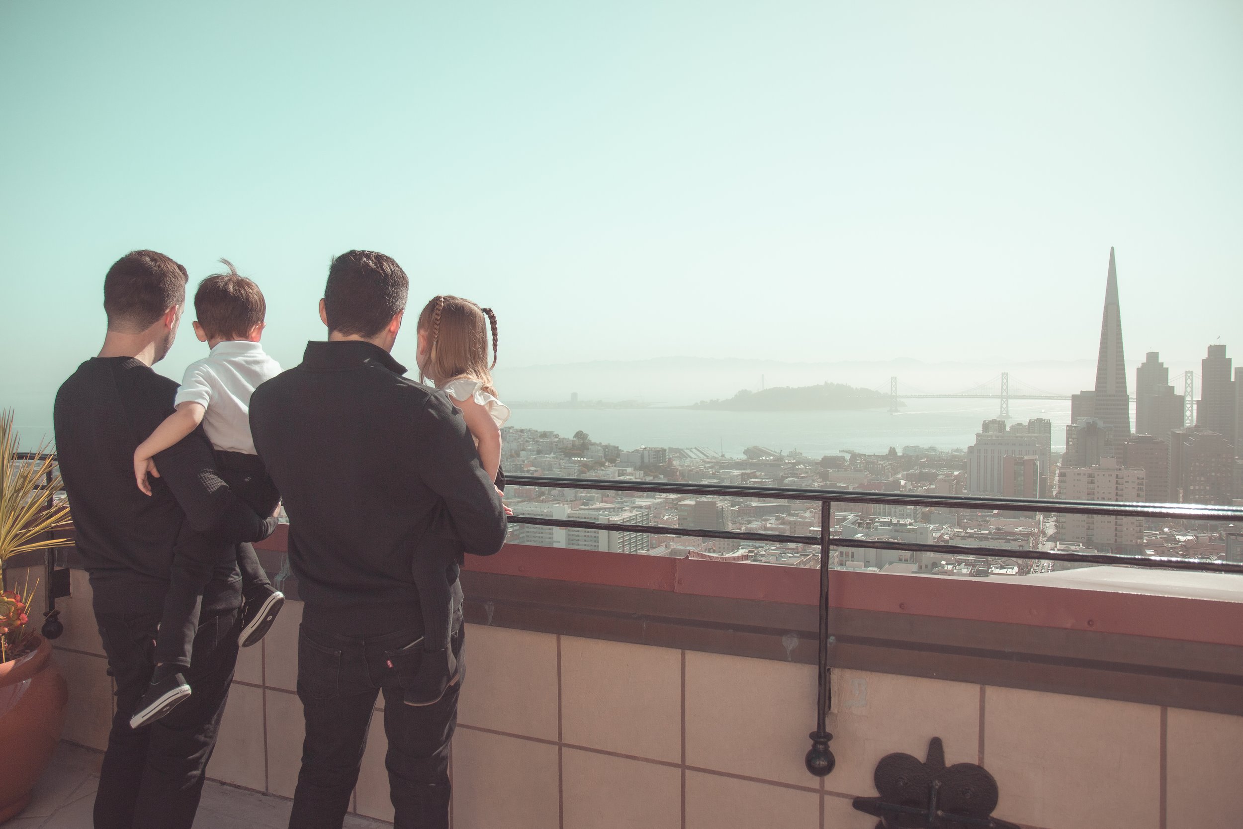 Four people, two men, a young boy, and a girl, stand on a balcony overlooking a city skyline with tall buildings and a bridge over a body of water.