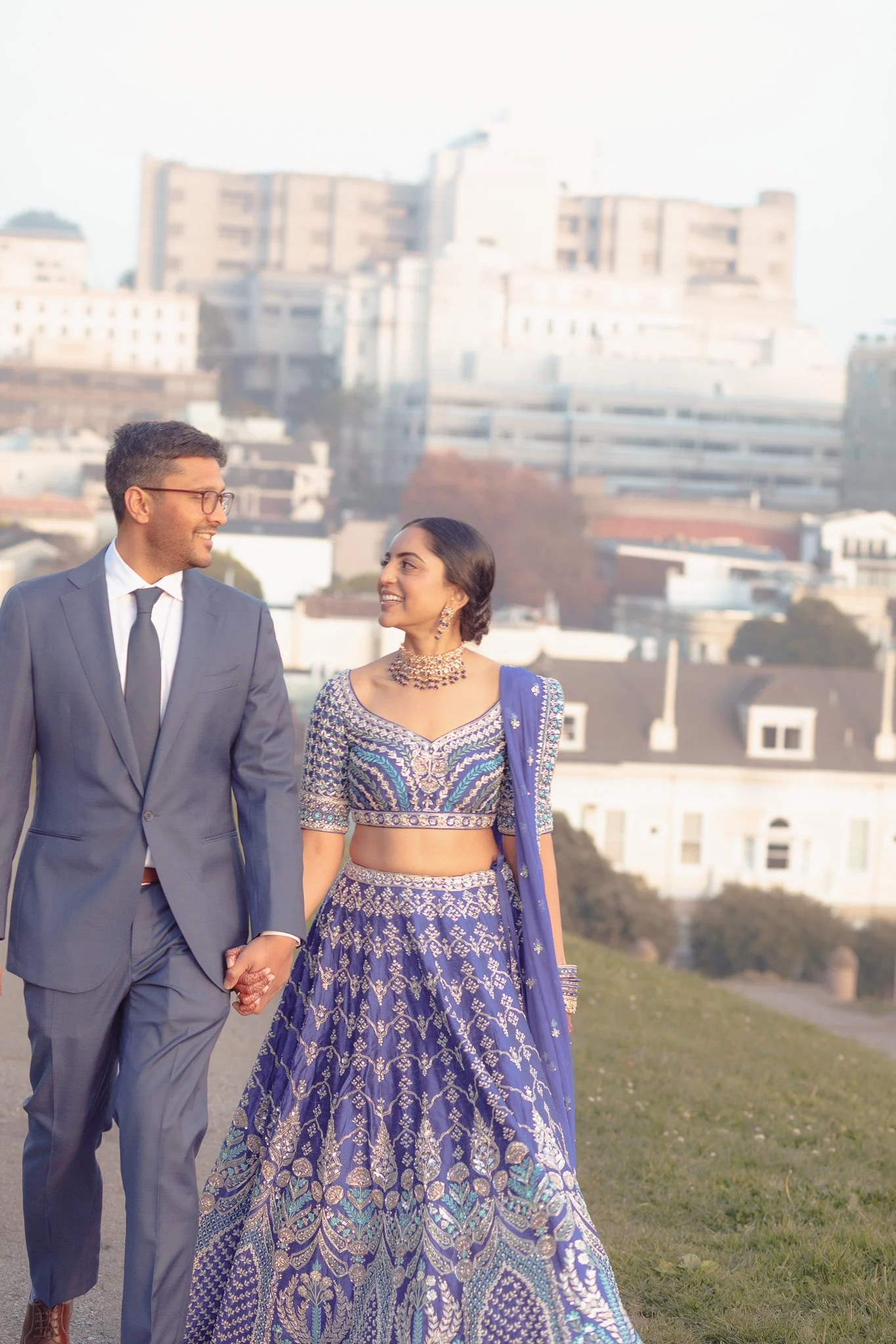 A couple walking hand in hand outdoors in formal attire, with the man in a suit and the woman in a blue traditional Indian outfit, smiling at each other, with city buildings in the background.
