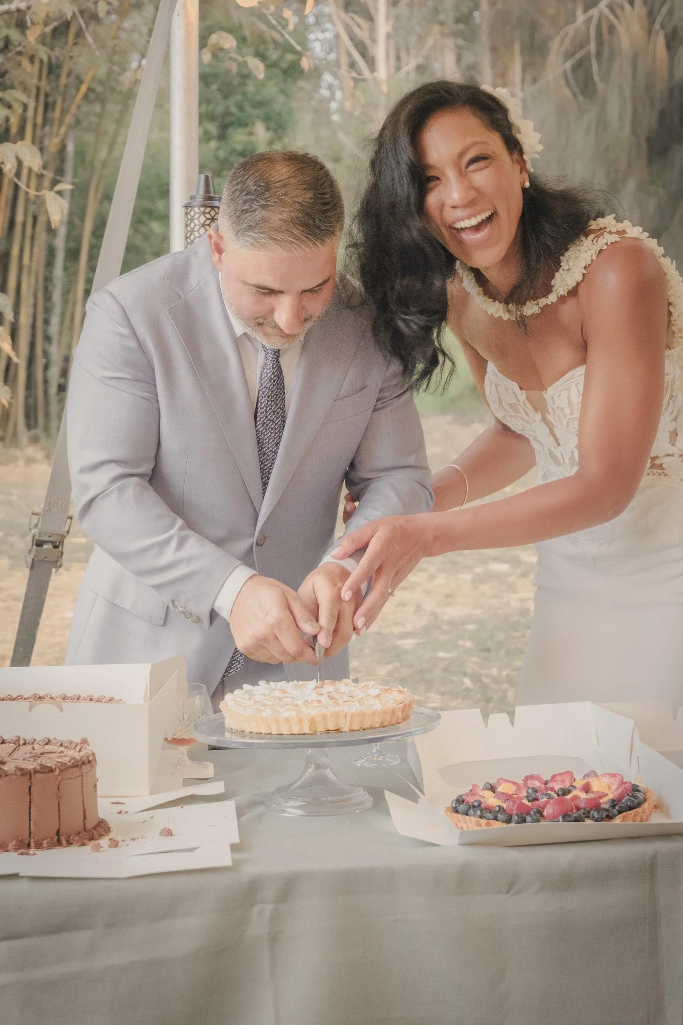 A bride and groom in wedding attire cutting a cake together at an outdoor reception, smiling and happy.
