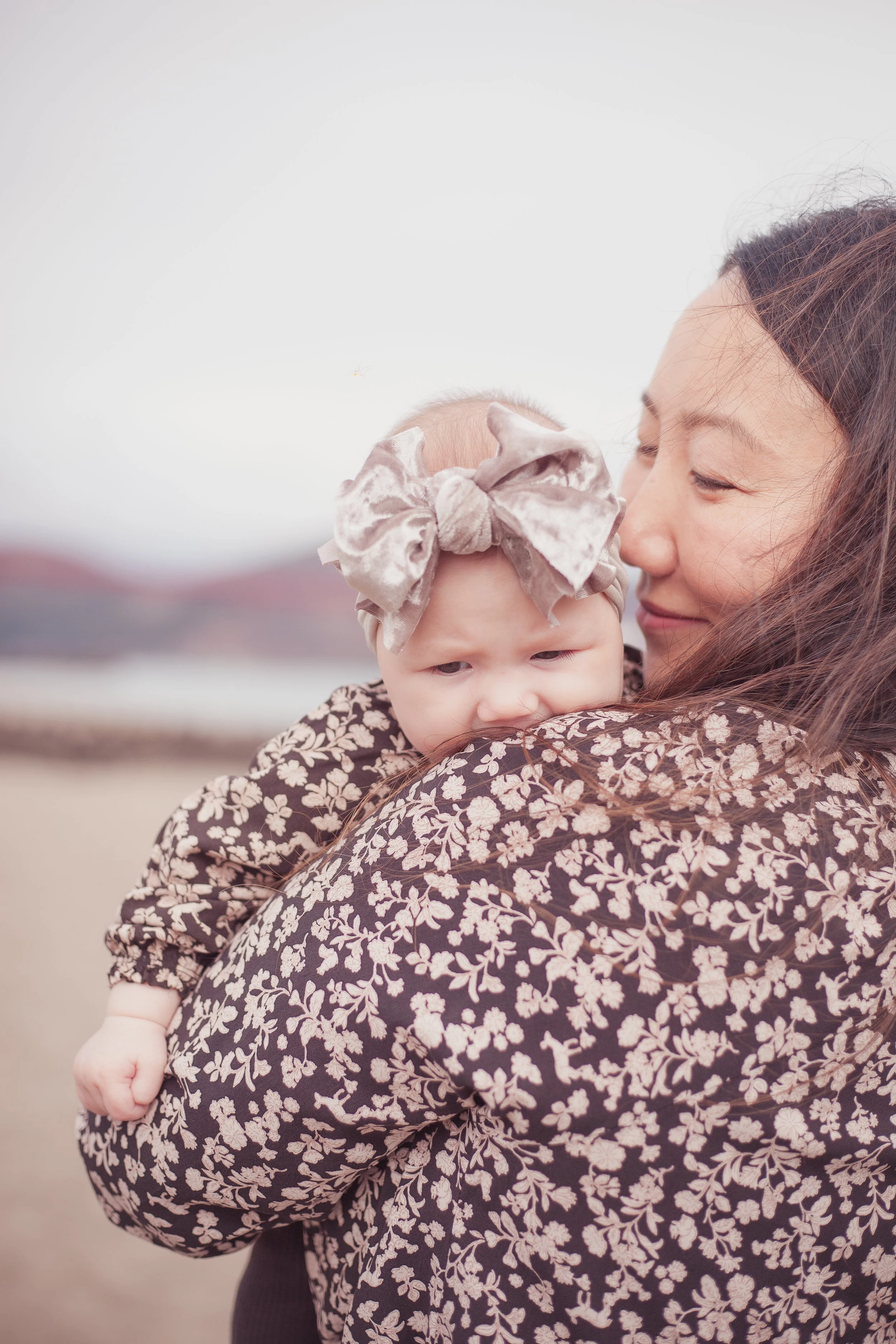 A woman holding a baby girl at the beach, with a blurred background of water and mountains.
