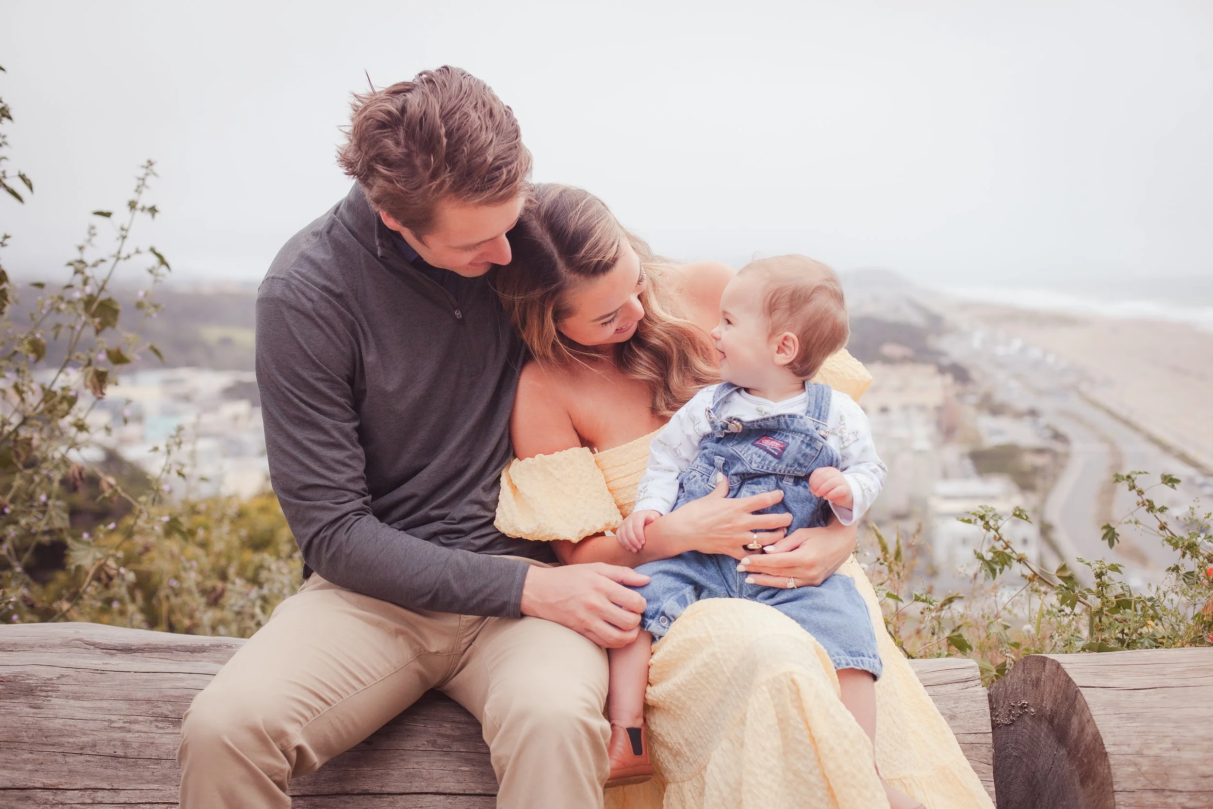 A family of three sitting on a wooden bench outdoors, with the mother holding a young child while the father leans in towards them, with a blurred coastal town and ocean in the background.
