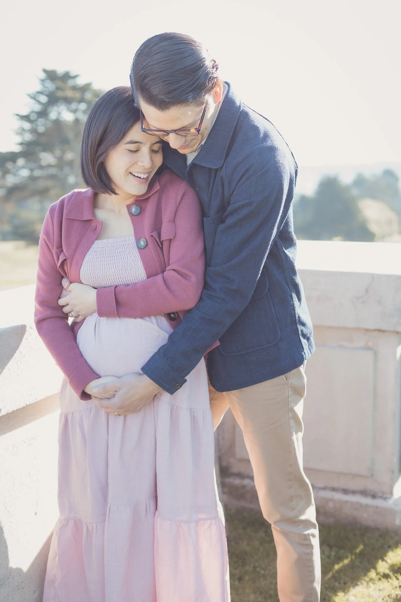 A man and a pregnant woman sharing a joyful moment outdoors, with the man kissing her forehead and both smiling.