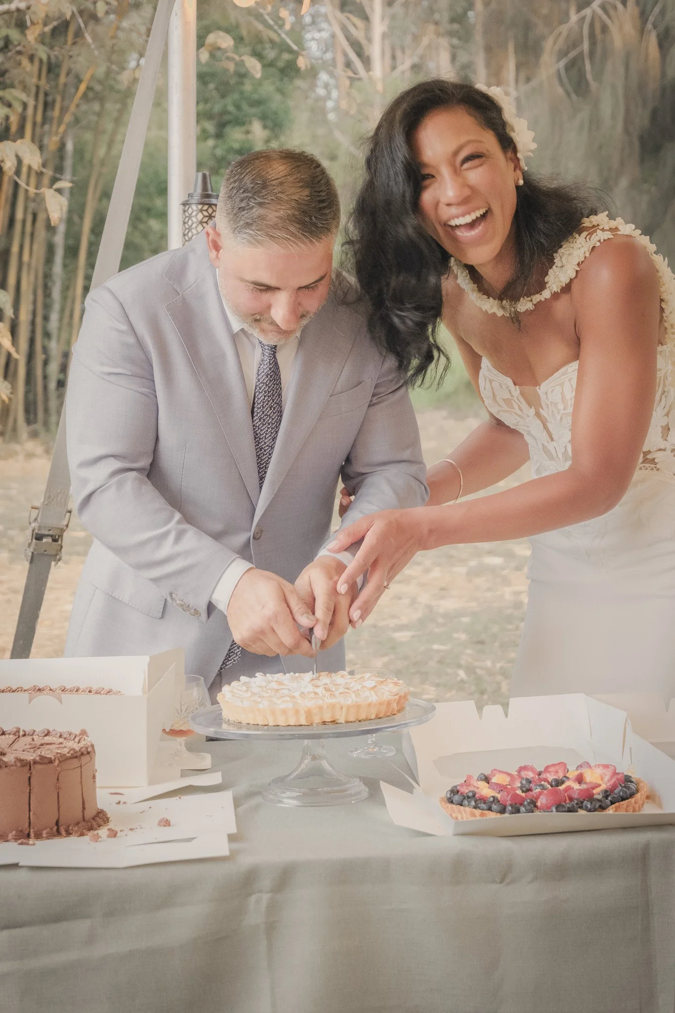 A couple cutting a wedding cake together at an outdoor wedding reception with other desserts on the table.