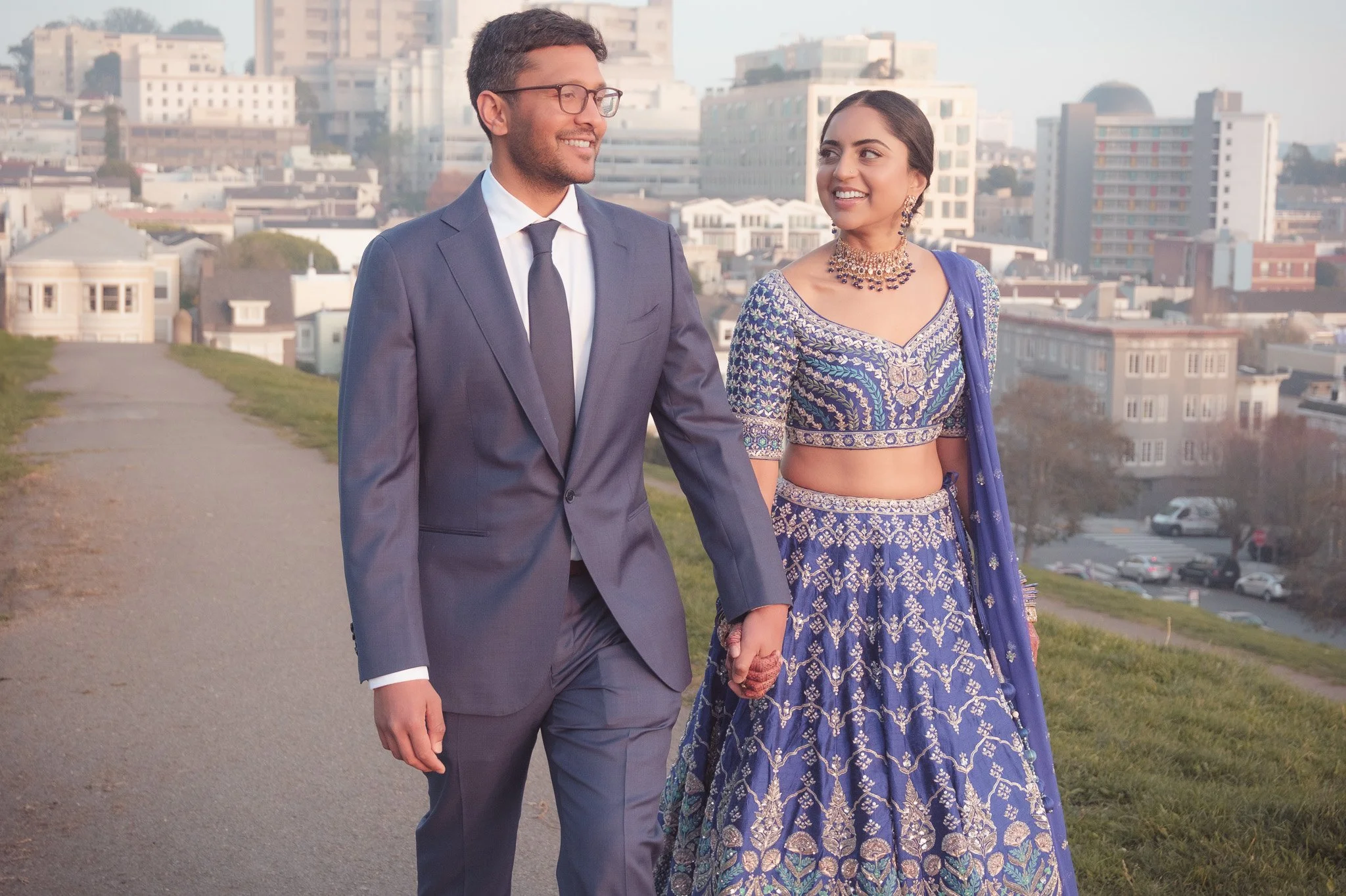 A couple walking hand in hand in a city park at sunset, the man in a gray suit and the woman in a traditional blue Indian outfit, smiling at each other.