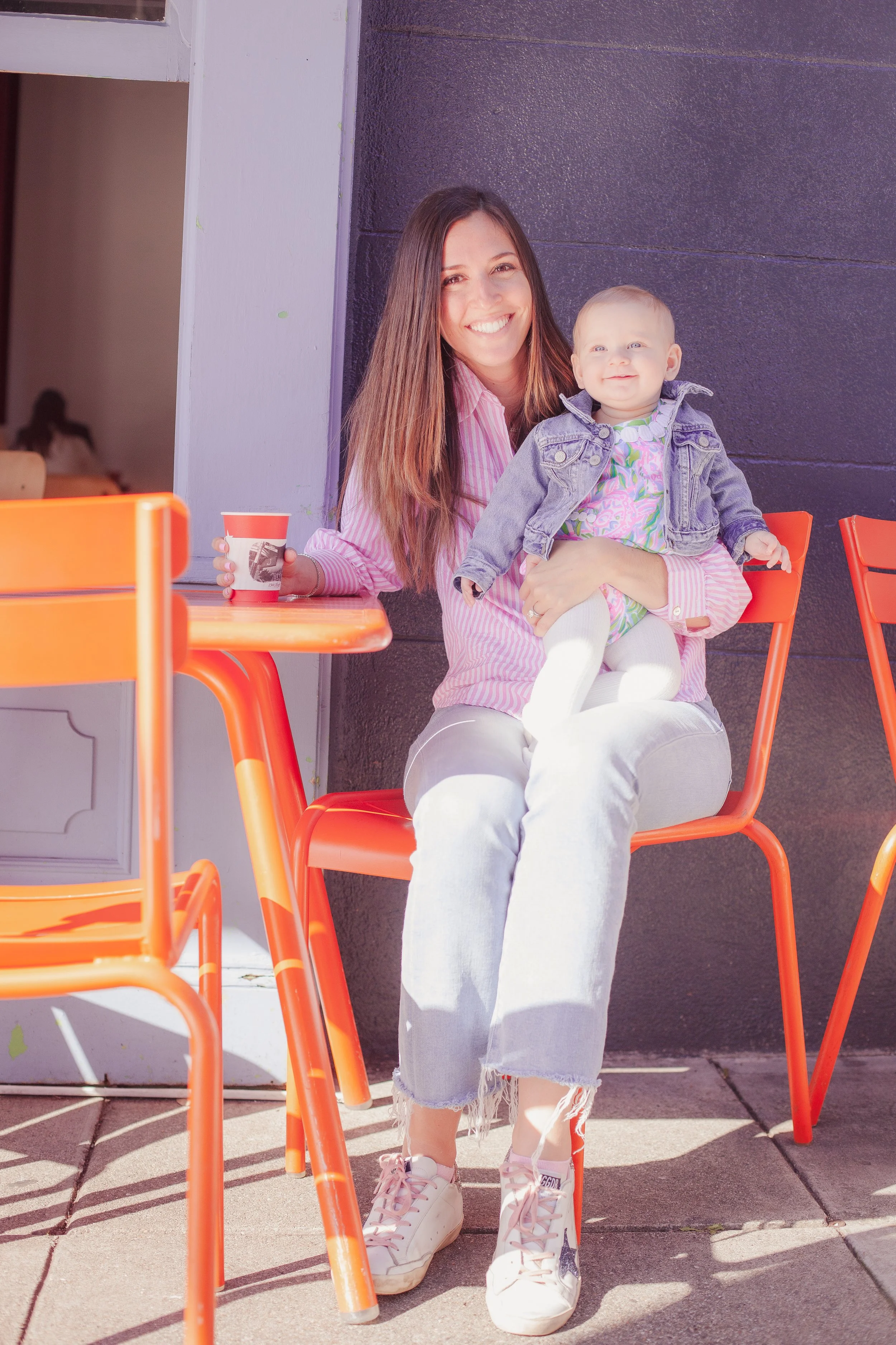 A woman sitting on an orange chair outside a cafe, smiling and holding a young girl on her lap. The girl is wearing a denim jacket and a colorful dress. The woman is dressed in a pink striped shirt, white jeans, and sneakers, with an orange table and