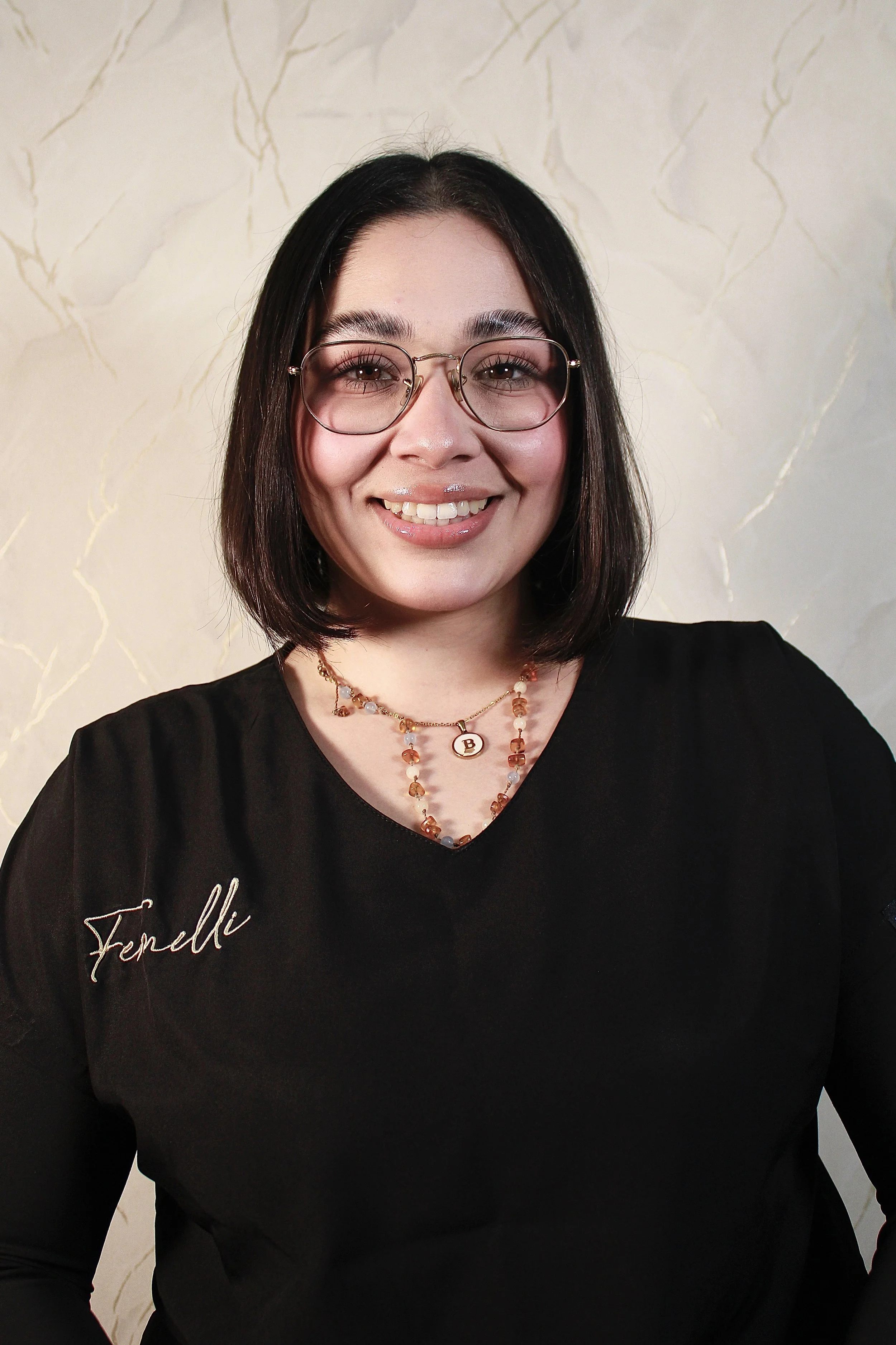 A woman with long dark hair wearing glasses, a black top with 'Fendli' embroidered on it, layered gold necklaces, and a white smartwatch is sitting against a light cream-colored wall with a subtle marble pattern.