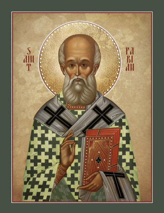 An icon-style depiction of a saint with a halo, gray beard, and gold background, holding a red book and making a gesture with his right hand. The saint is dressed in green and gray robes with black crosses.