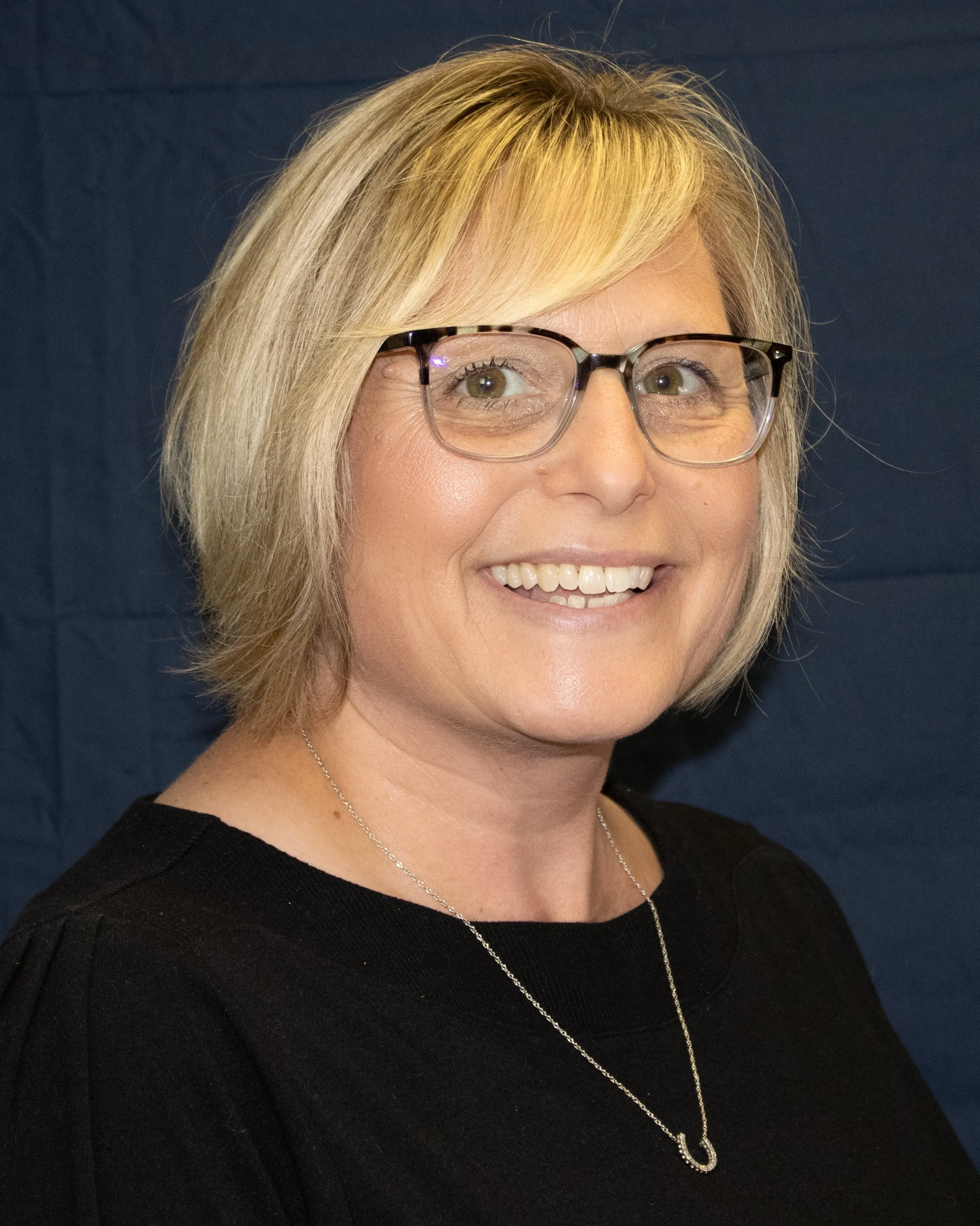 A smiling woman with short blonde hair, glasses, wearing a black top and a silver necklace, standing against a dark background.