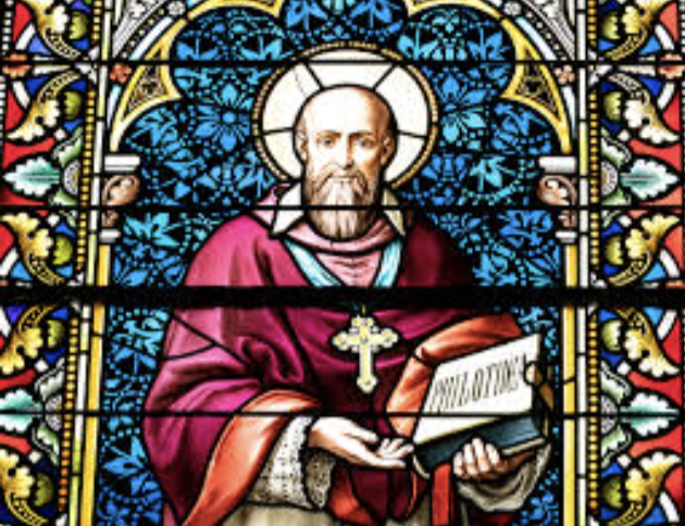 Stained glass window depicting a religious figure with a halo, holding a book and a staff with a cross, surrounded by colorful floral patterns.