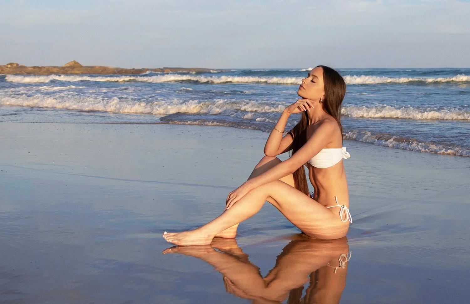 Female model photoshoot on location at Curl Curl beach Sydney wearing a white bikini sitting on the waters edge at sunset