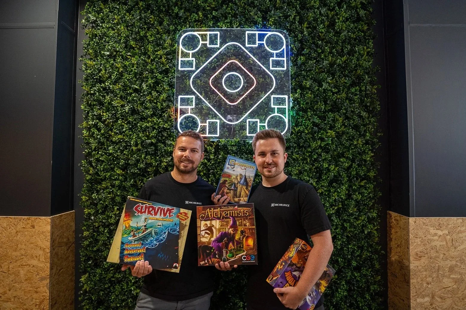 Two men holding board games in front of a green wall and illuminated dice sign