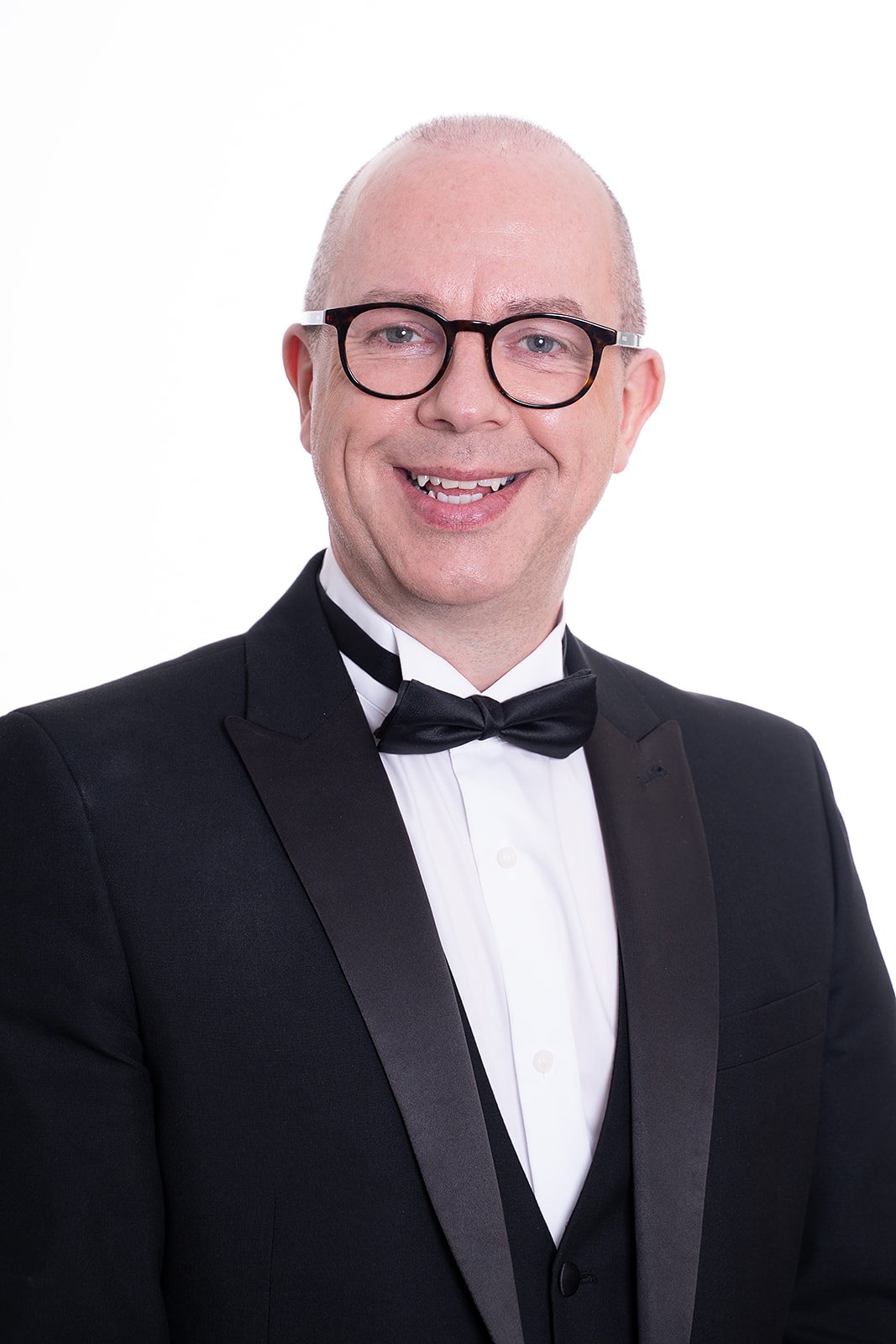 A smiling man wearing glasses, a black tuxedo, white shirt, and black bow tie, posing against a white background.