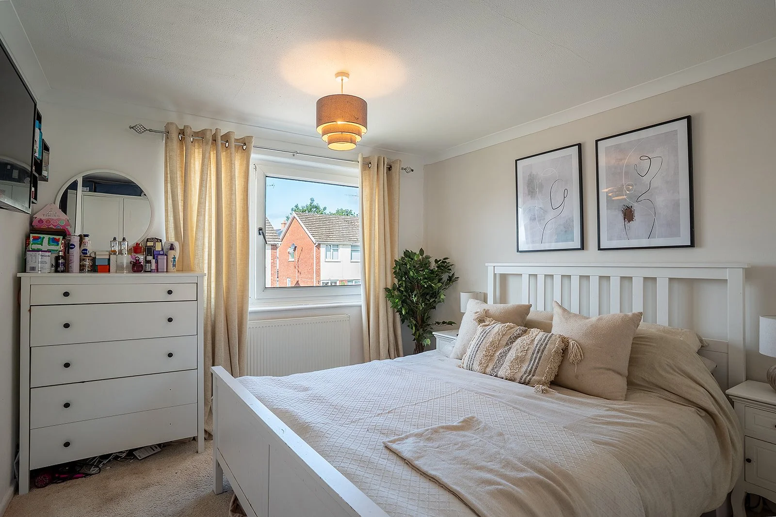 Bright neutral bedroom with a white bed, cream curtains, framed wall art and a chest of drawers.