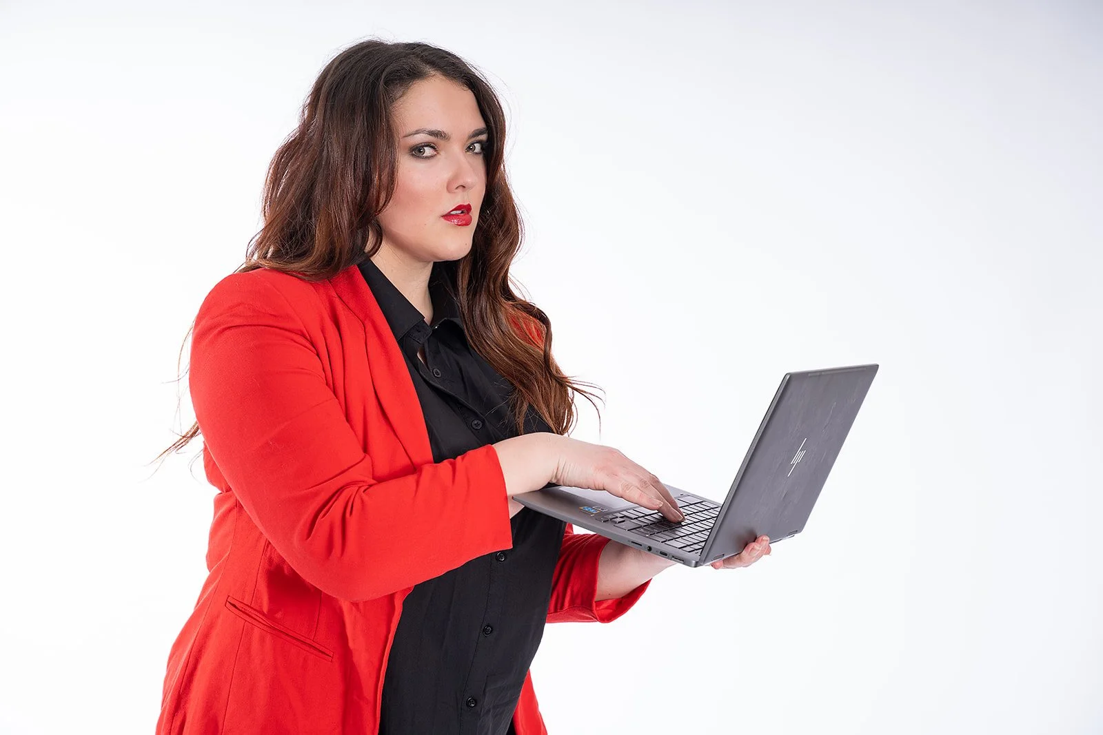 Woman with long brown hair wearing a red blazer and black shirt holding a laptop and looking at the camera.