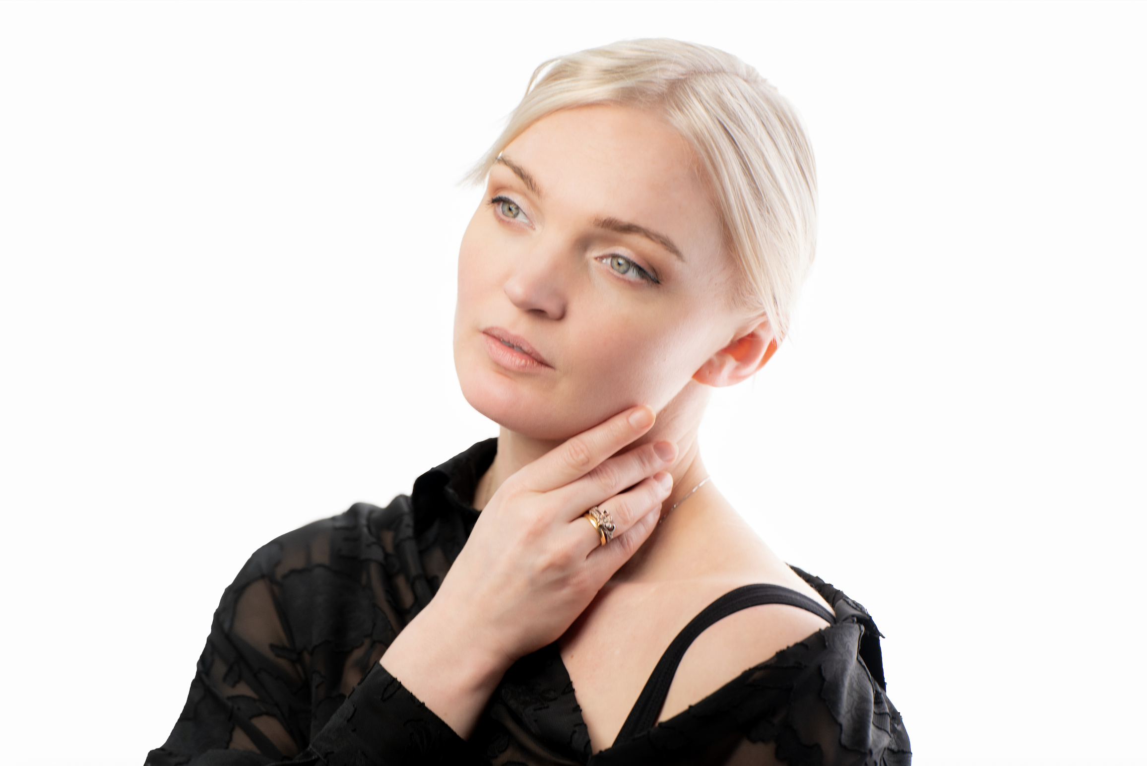 A young woman with blonde hair and blue eyes touching her neck, wearing a black lace top and a ring, against a white background.