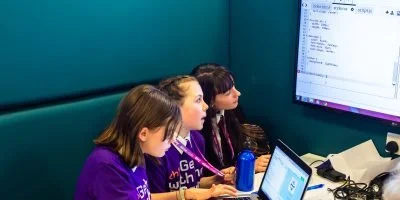 Three young girls working together on laptops in a classroom setting, focused on a large monitor displaying code or a program.