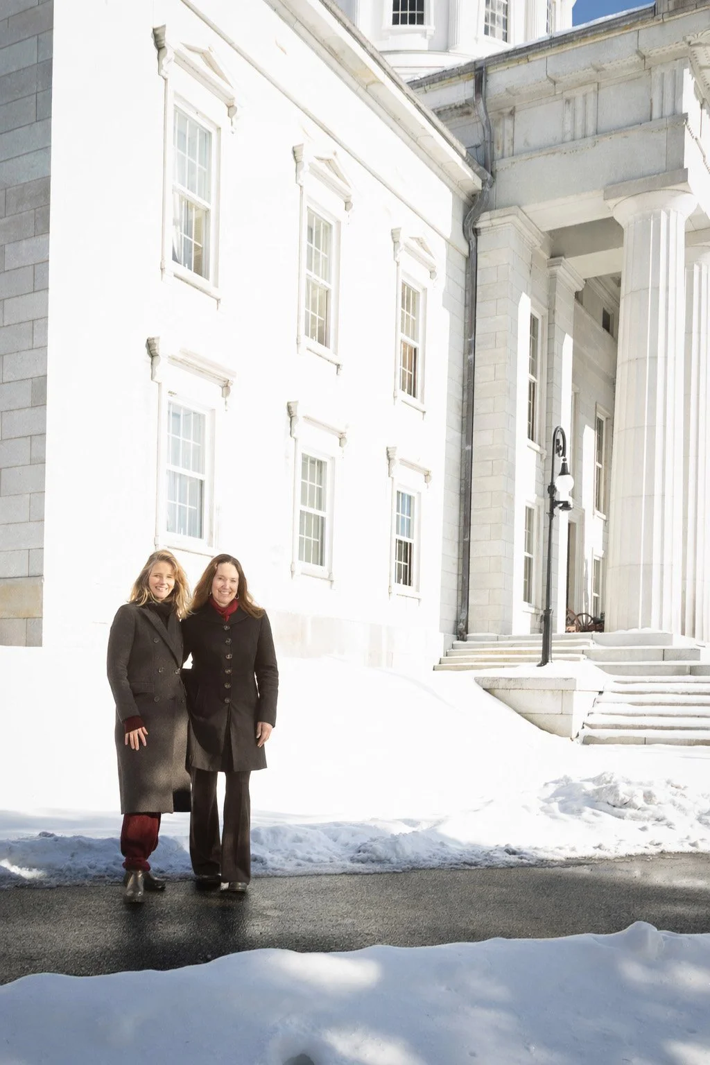 Two women standing outside on snow-covered ground in front of a large, white, historic building with columns and multiple windows.