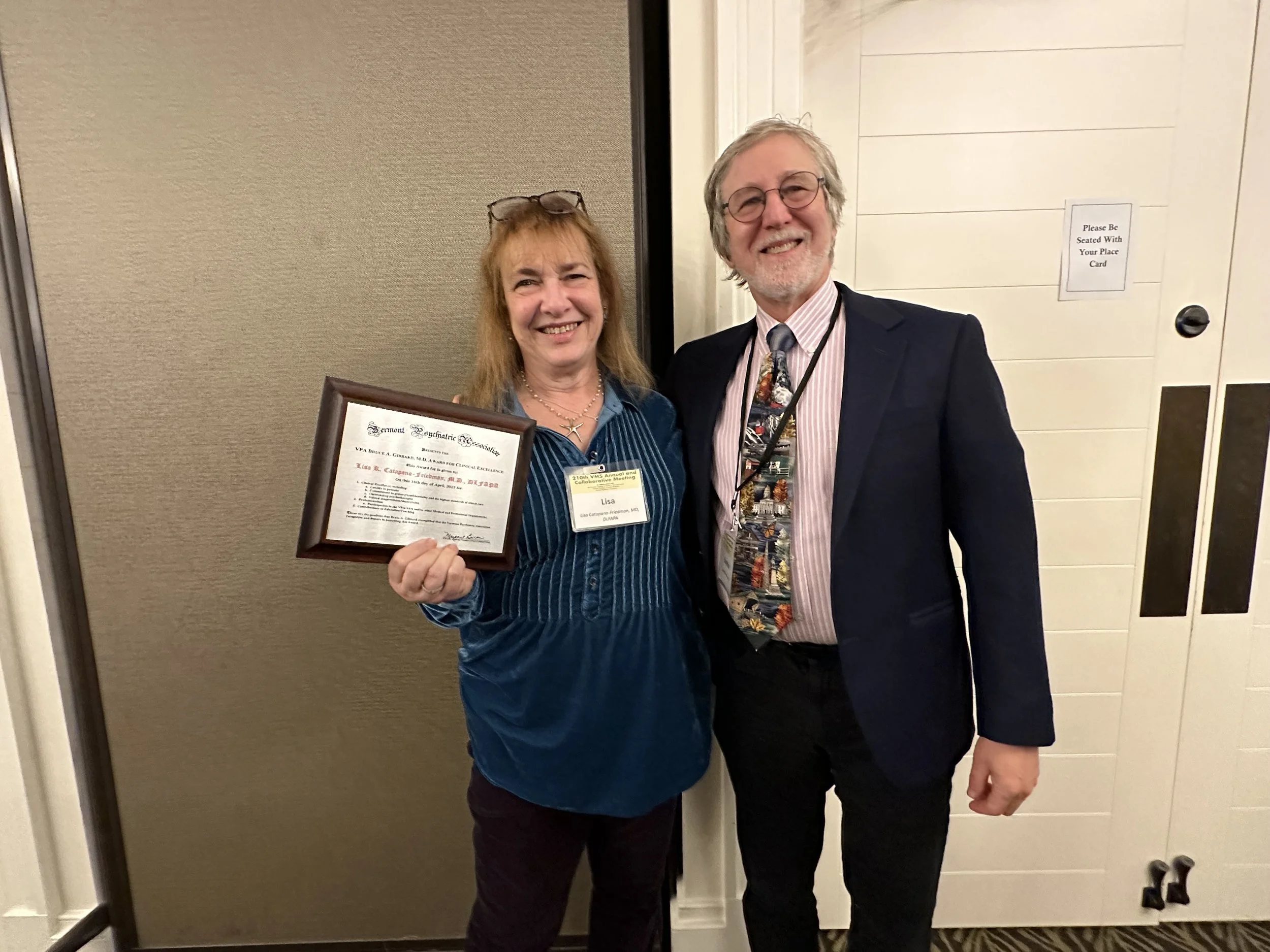 A smiling woman holding a framed certificate standing next to a smiling man in a suit with a colorful tie, in an indoor setting.