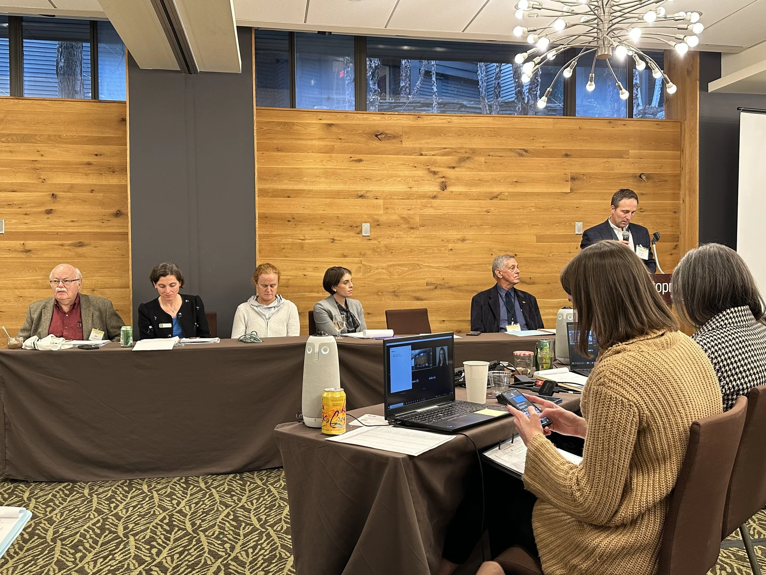 People sitting at a conference table with a speaker standing at a podium, addressing the audience in a room with wood-paneled walls and a chandelier overhead. Some attendees are using laptops and phones.
