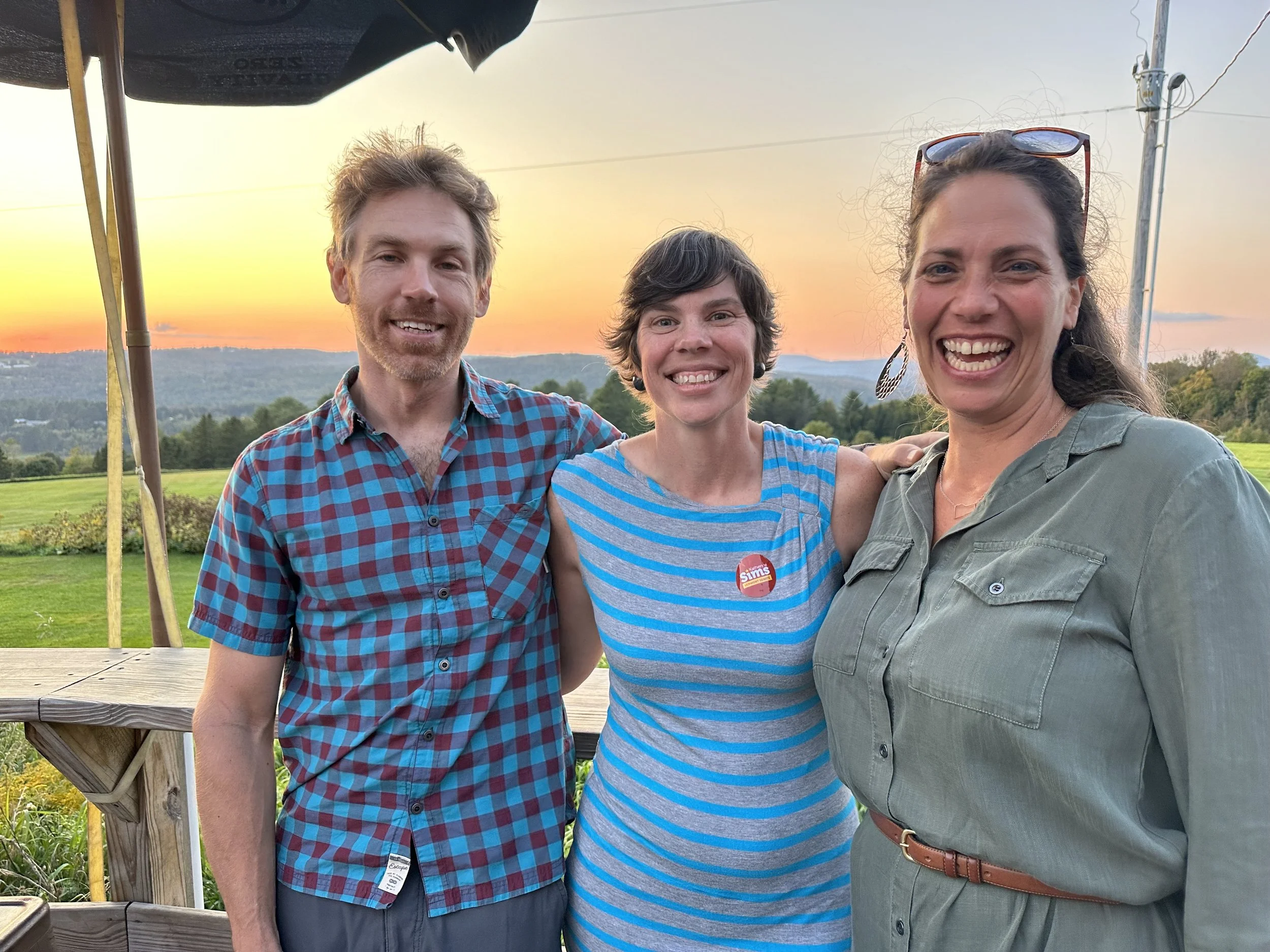 Three people smiling and posing outdoors during sunset, standing on a wooden deck with green fields and hills in the background. The person in the middle is wearing a striped blue and gray dress with a campaign button, the person on the right is wear