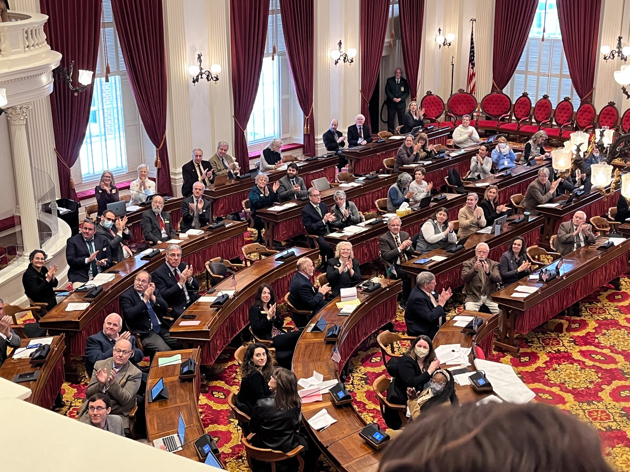 A formal assembly in a legislative chamber with officials seated at curved desks, many clapping. The room features tall windows with red curtains, ornate chandeliers, and an American flag. People are dressed in business attire, with some wearing mask