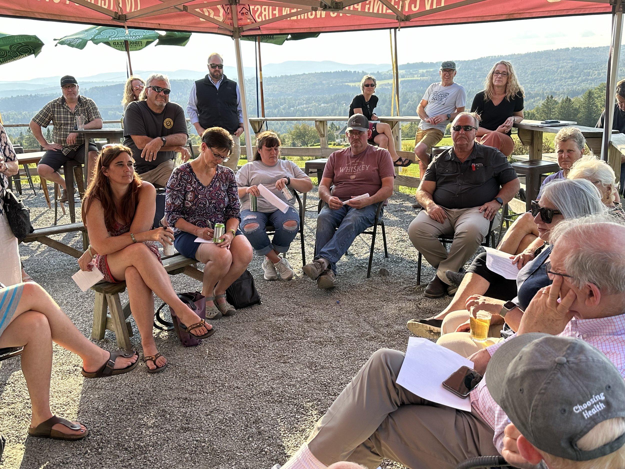 A group of people gathered outdoors under a canopy, sitting and standing on benches and chairs, with scenic mountains in the background. Some are holding papers and drinks, participating in a discussion or meeting.