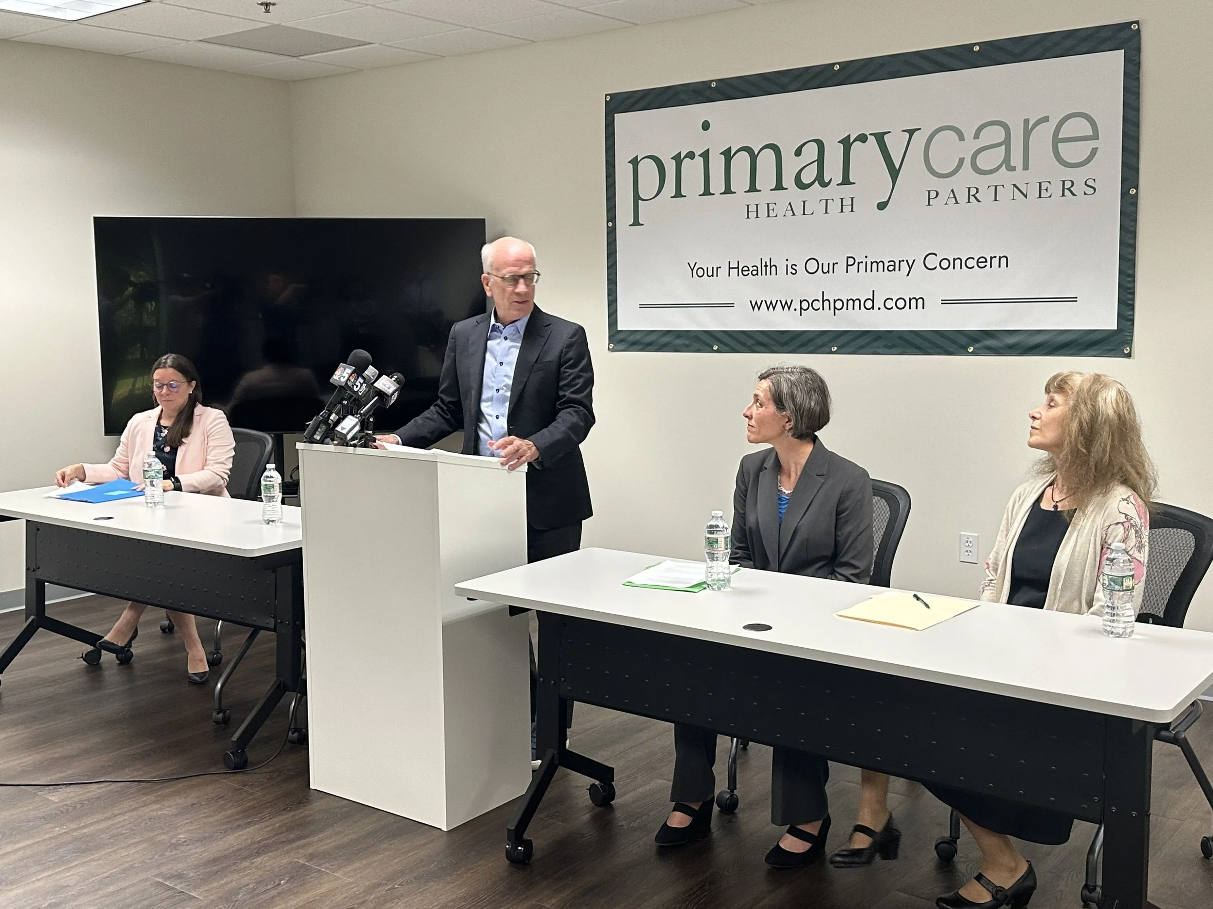 A man in a suit speaking at a podium during a press conference, with three women seated at tables on either side, in a room with a large sign for Primary Care Health Partners on the wall.
