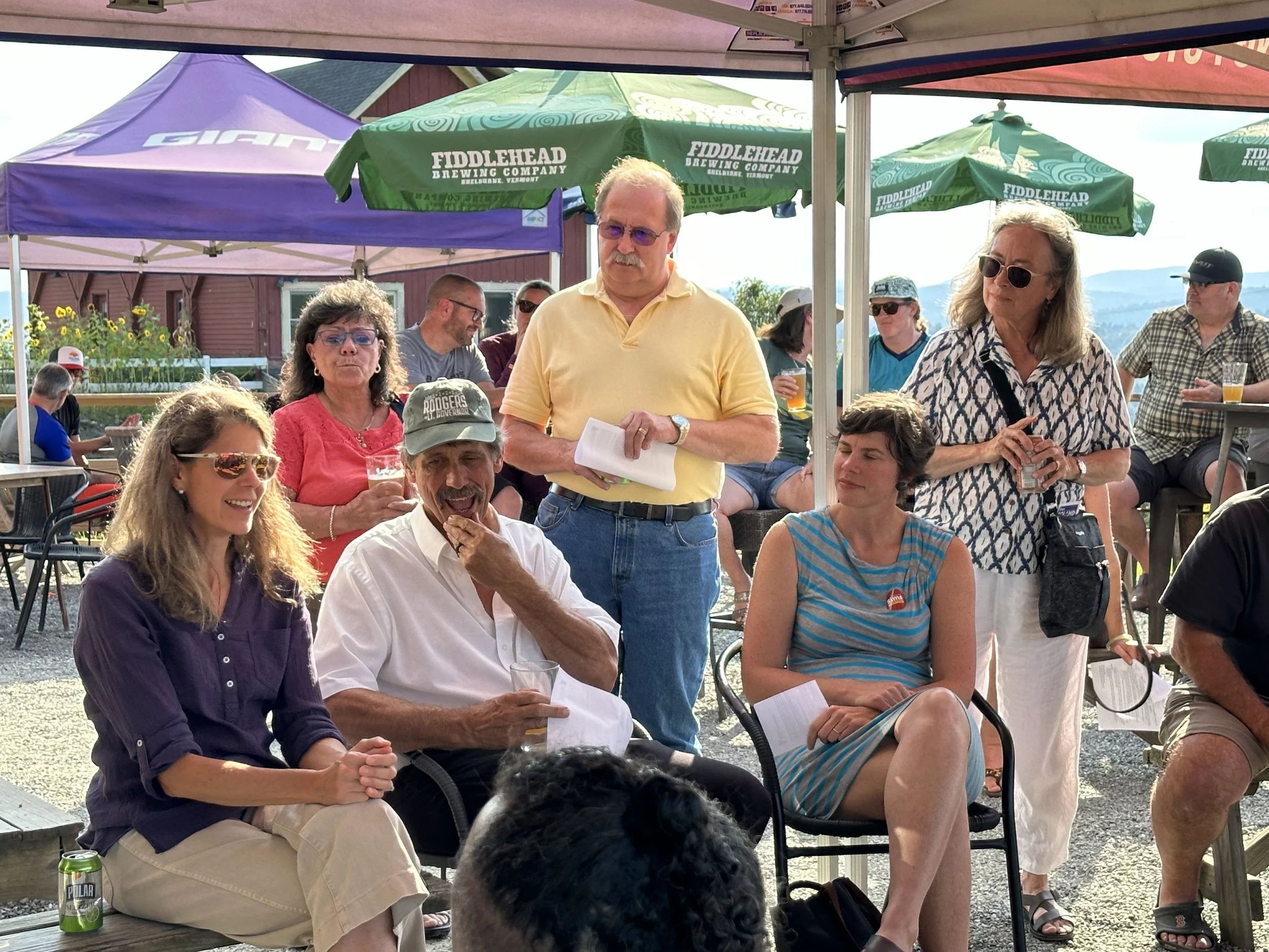 Group of people at an outdoor gathering under tents, sitting and standing, holding drinks and papers, with a rural background and trees.