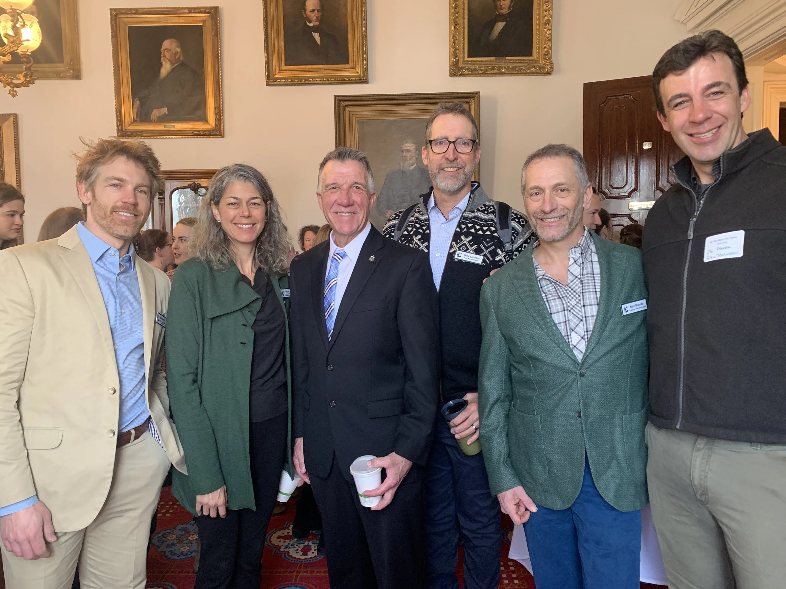 A group of six diverse professionals, four men and two women, standing together indoors at a formal event with framed portraits on the wall behind them. They are smiling at the camera and some are holding coffee cups.