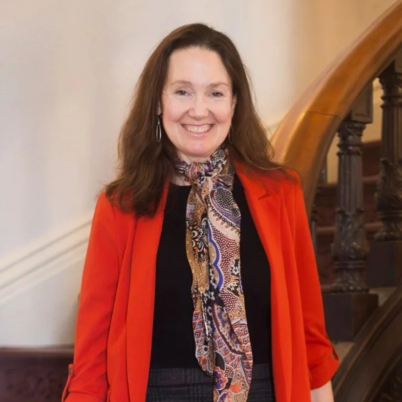 A woman with shoulder-length brown hair smiling, wearing a black top, an orange blazer, and a colorful patterned scarf, standing indoors near a wooden staircase.