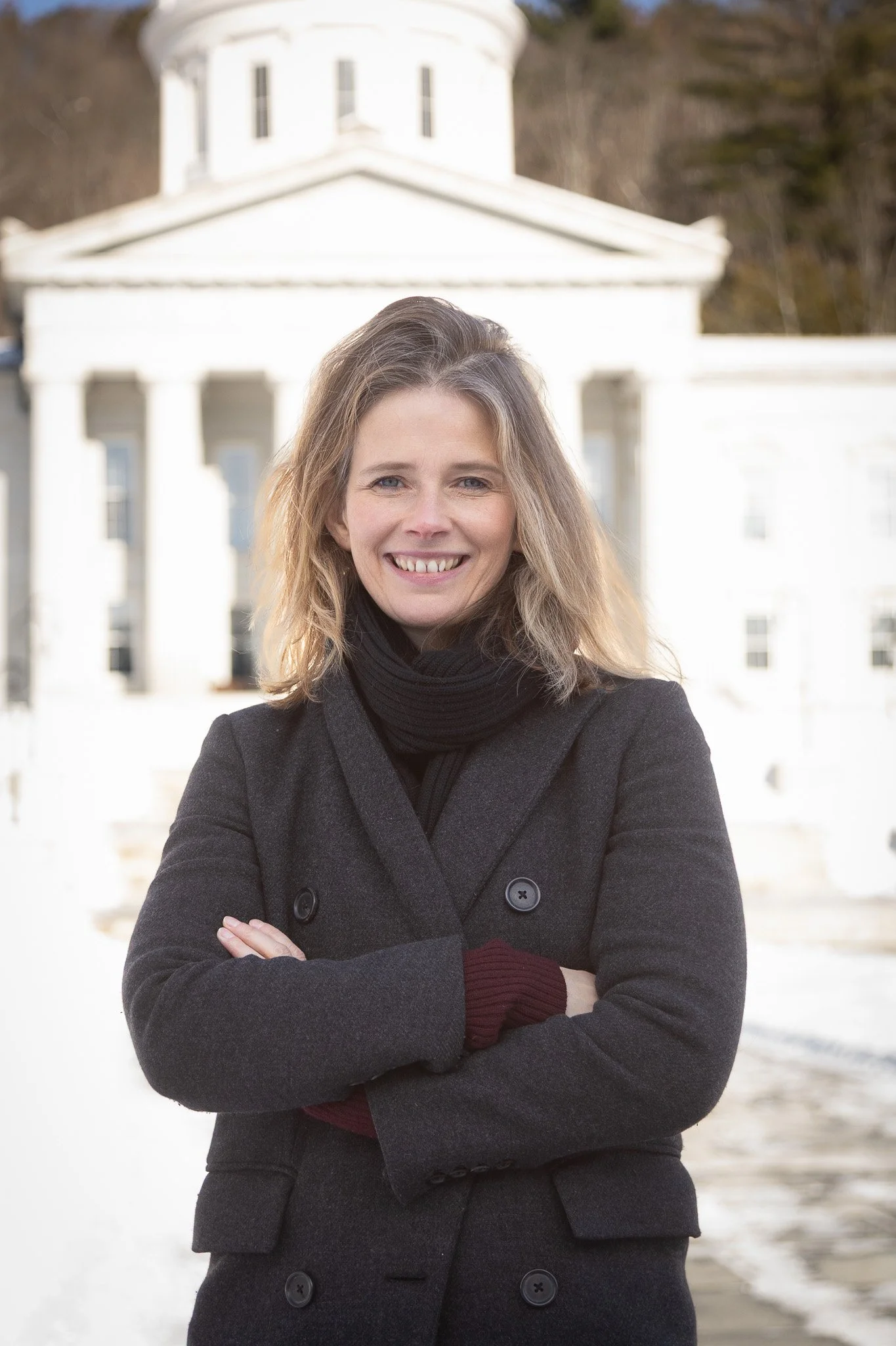 A woman with shoulder-length blonde hair, wearing a gray coat and black scarf, standing with arms crossed in front of the White House.