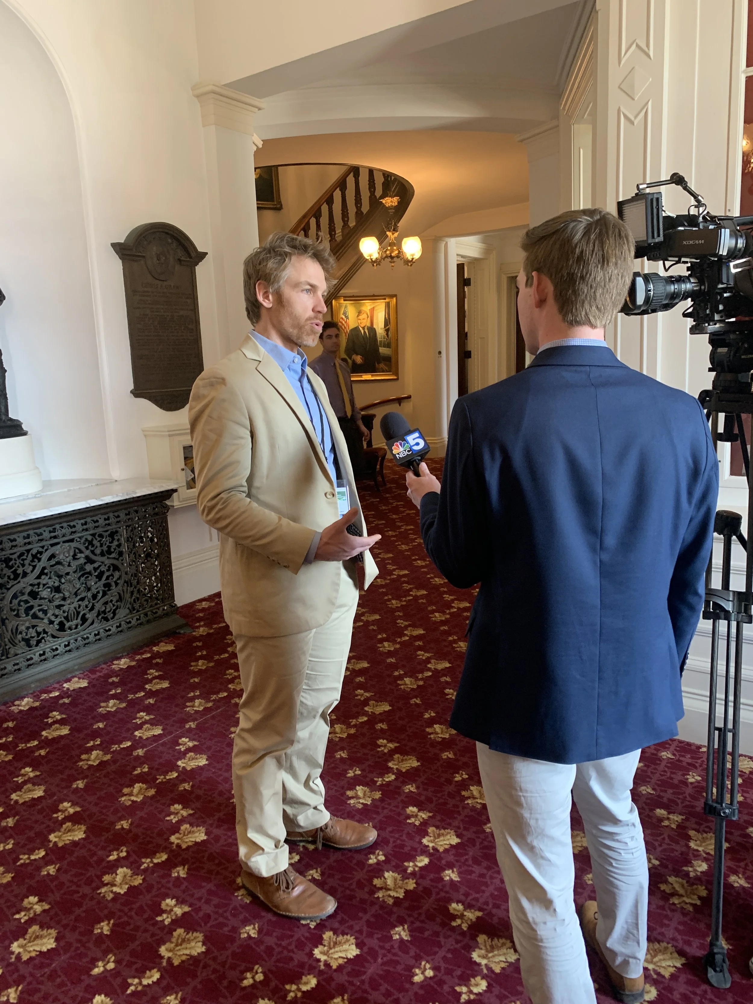 A man in a beige suit being interviewed by a reporter with a NBC 5 microphone in a hallway of an elegant building.