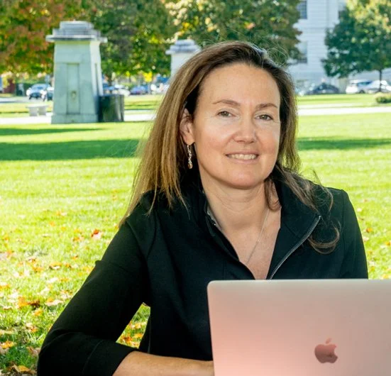 Woman sitting outdoors at a park with a laptop, smiling, with trees and park statues in the background.