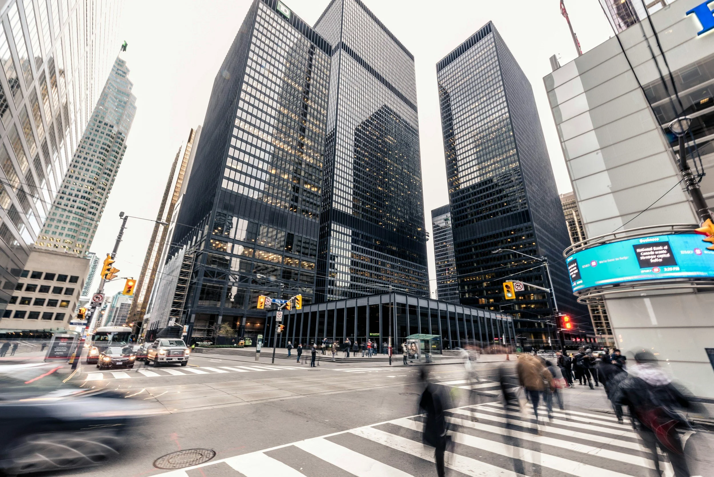 City intersection with tall glass office buildings, pedestrians crossing the street, cars in motion, traffic lights, and bright digital billboards.