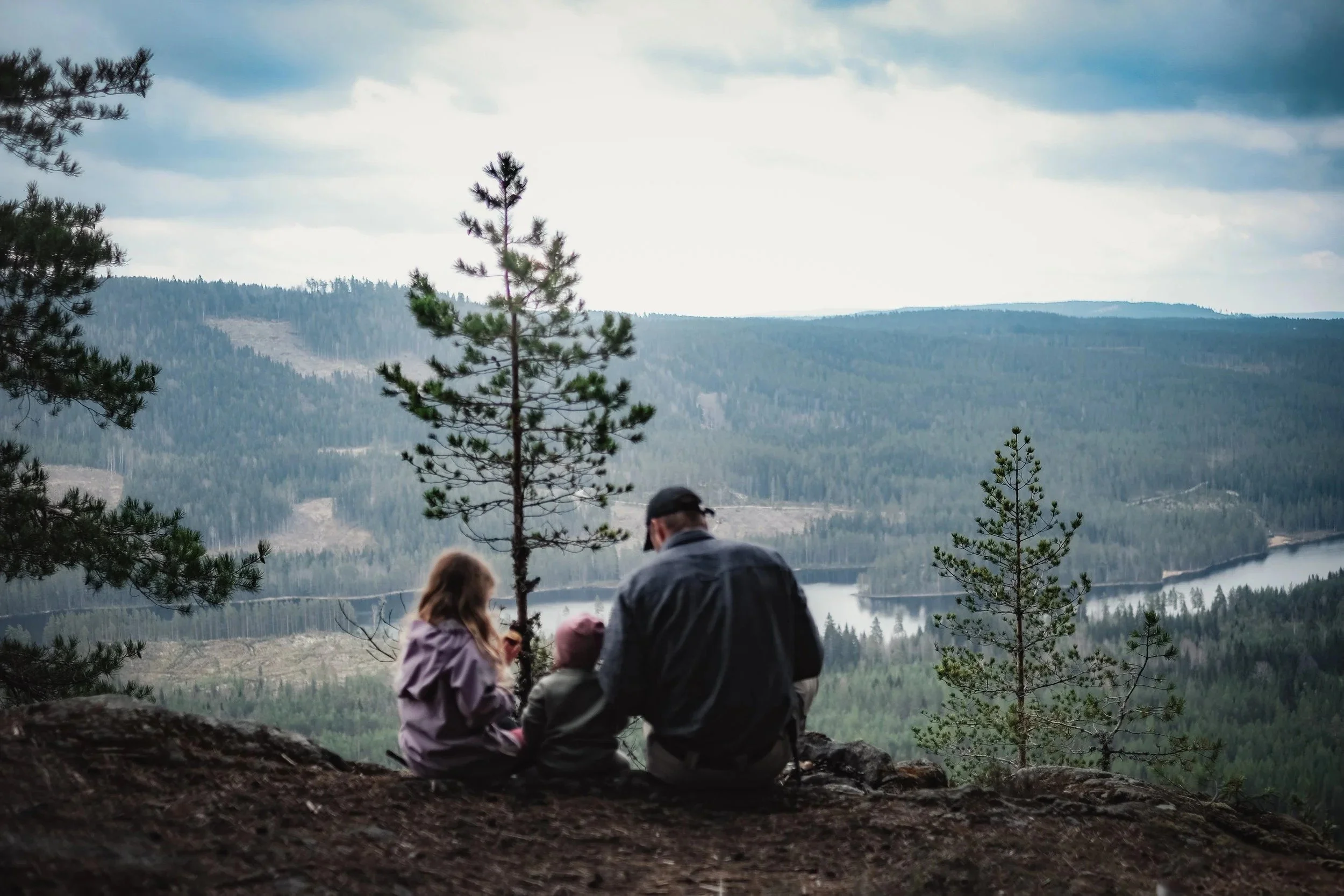 An adult and two children sitting on a viewpoint looking out over a vast forest landscape and a river.