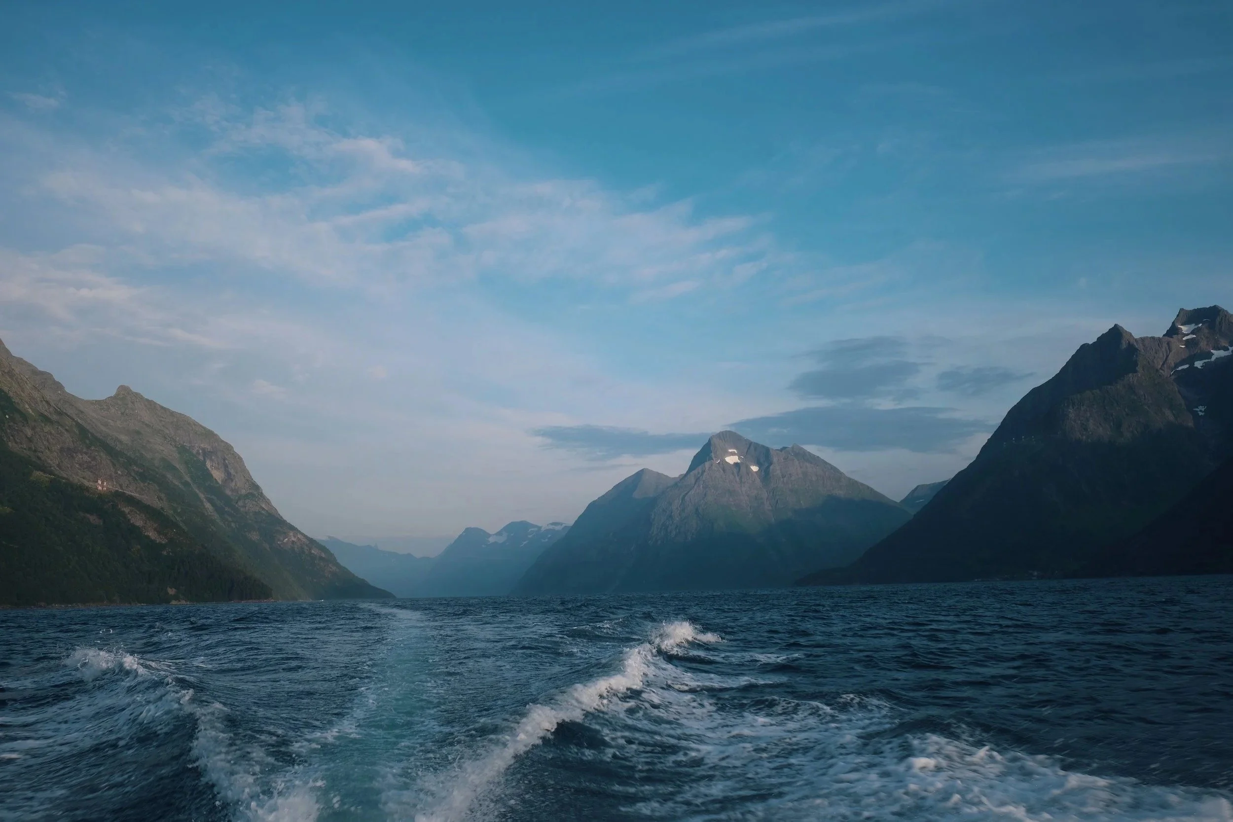 View from the back of a boat cruising through a deep fjord surrounded by steep mountains.