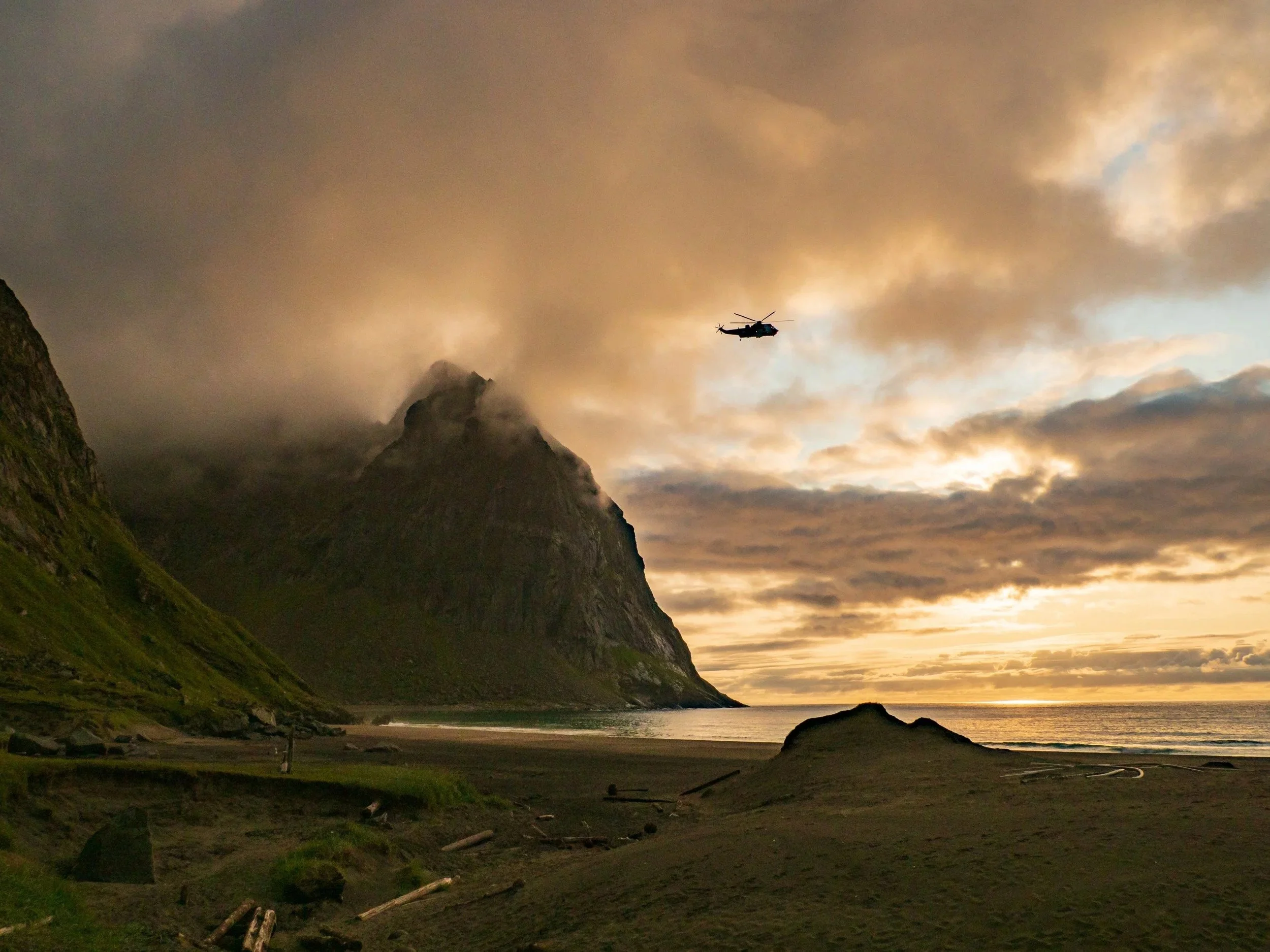 A helicopter flying over a coastal beach flanked by steep, misty mountains under a sunset sky.